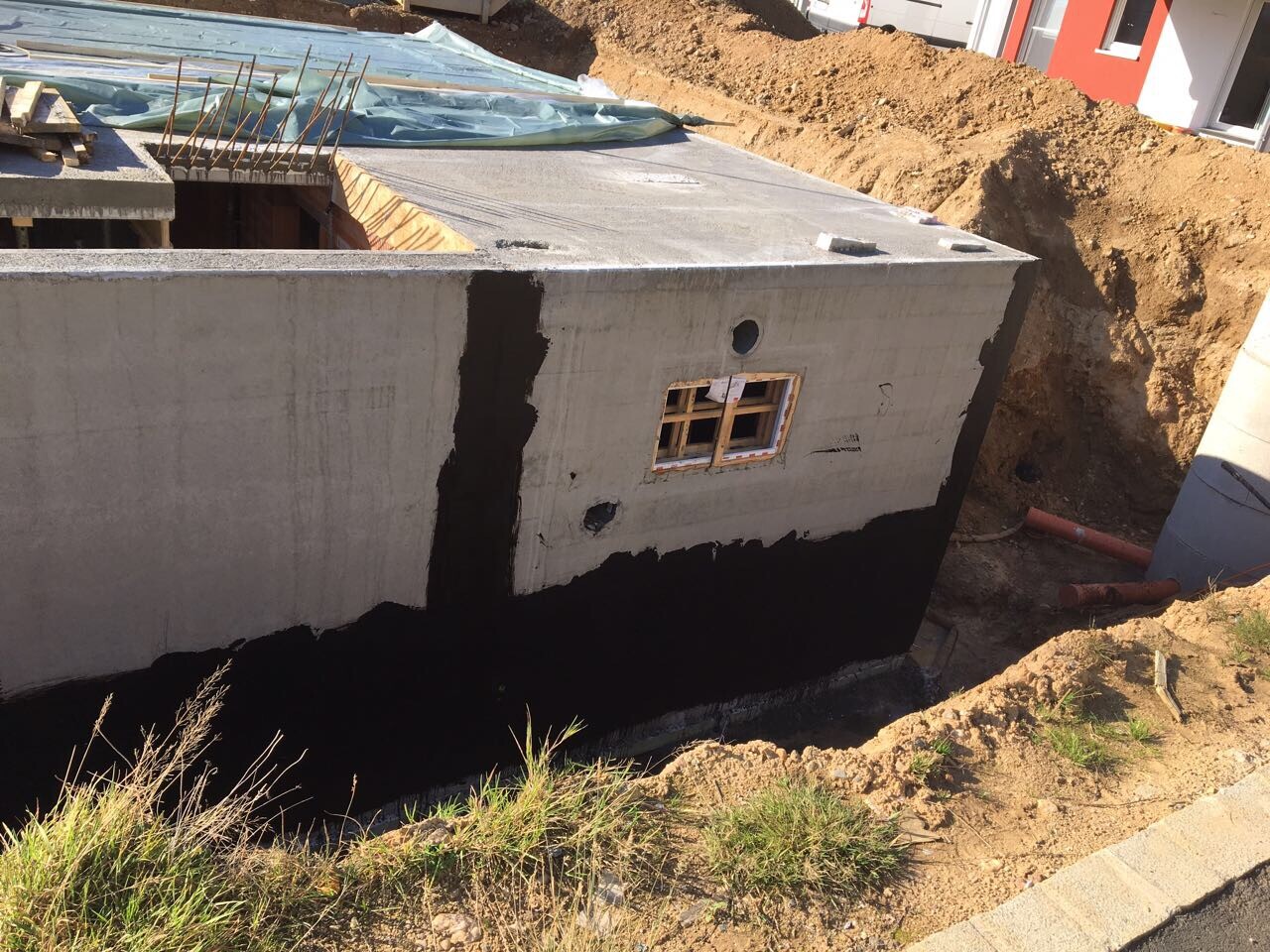 Betonfundament mit kleiner Fensteröffnung, dunkler Abdichtung und Erdarbeiten rundherum.