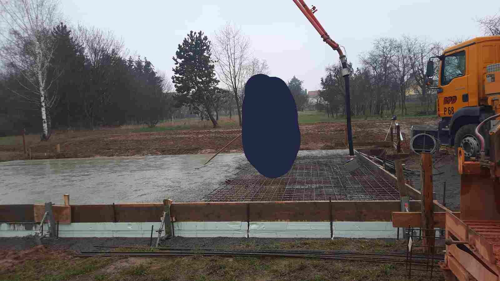 Baustelle mit Betonfundament, Bewehrungsstahlgitter, Betonpumpe und gelbem LKW; Bäume im Hintergrund.