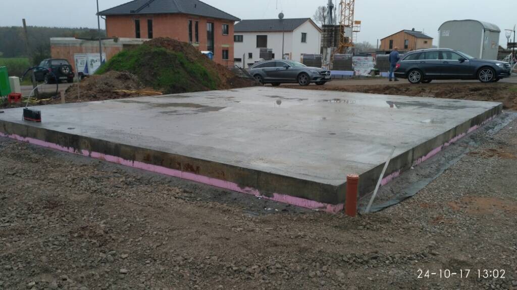Betonfundamentplatte auf Baustelle, pinke Dämmung entlang der Kante, Fahrzeuge im Hintergrund.