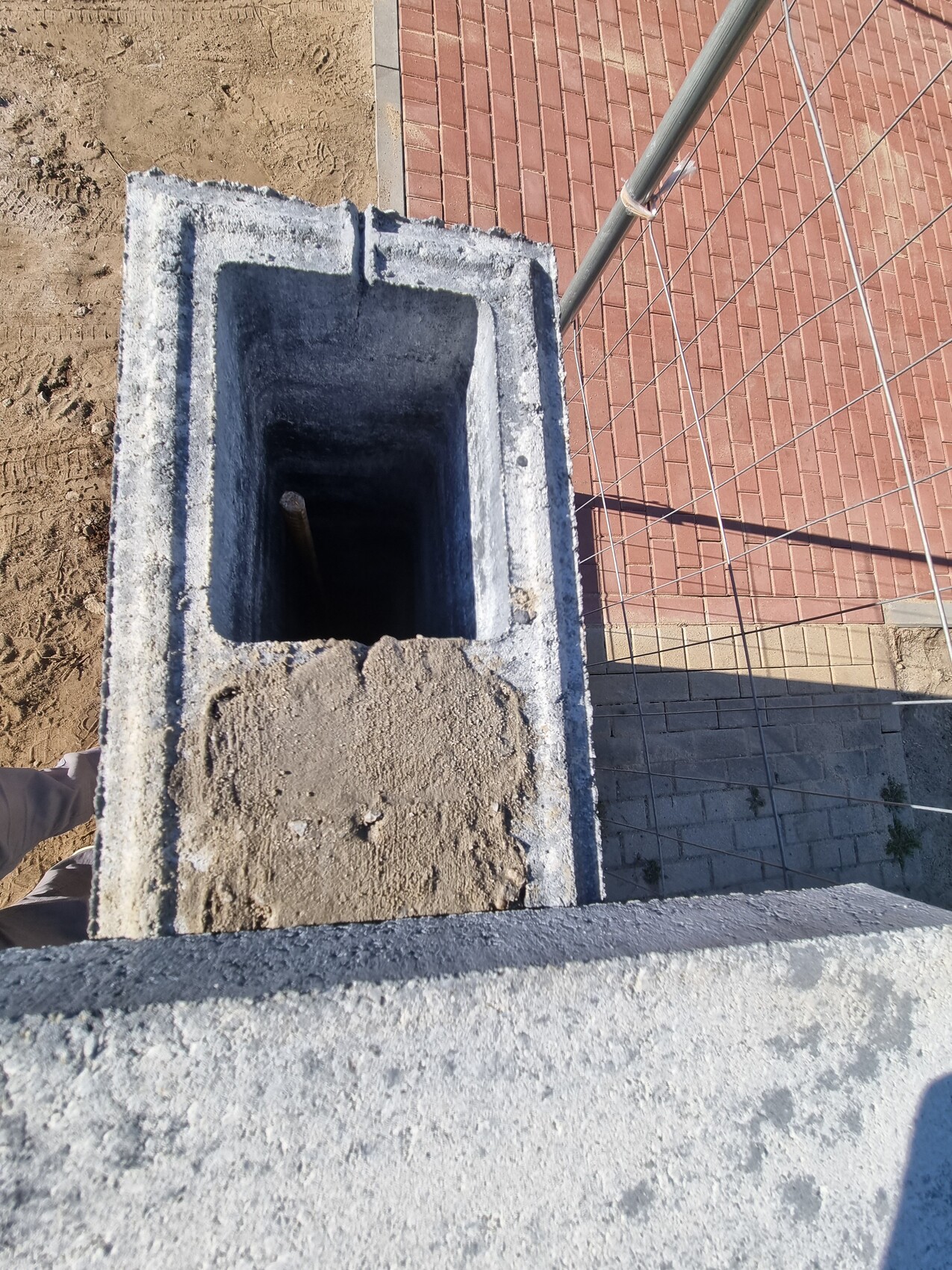 Beton-Schacht mit Innenraum, Blick von oben; Baustellenzaun und roter Pflasterweg.