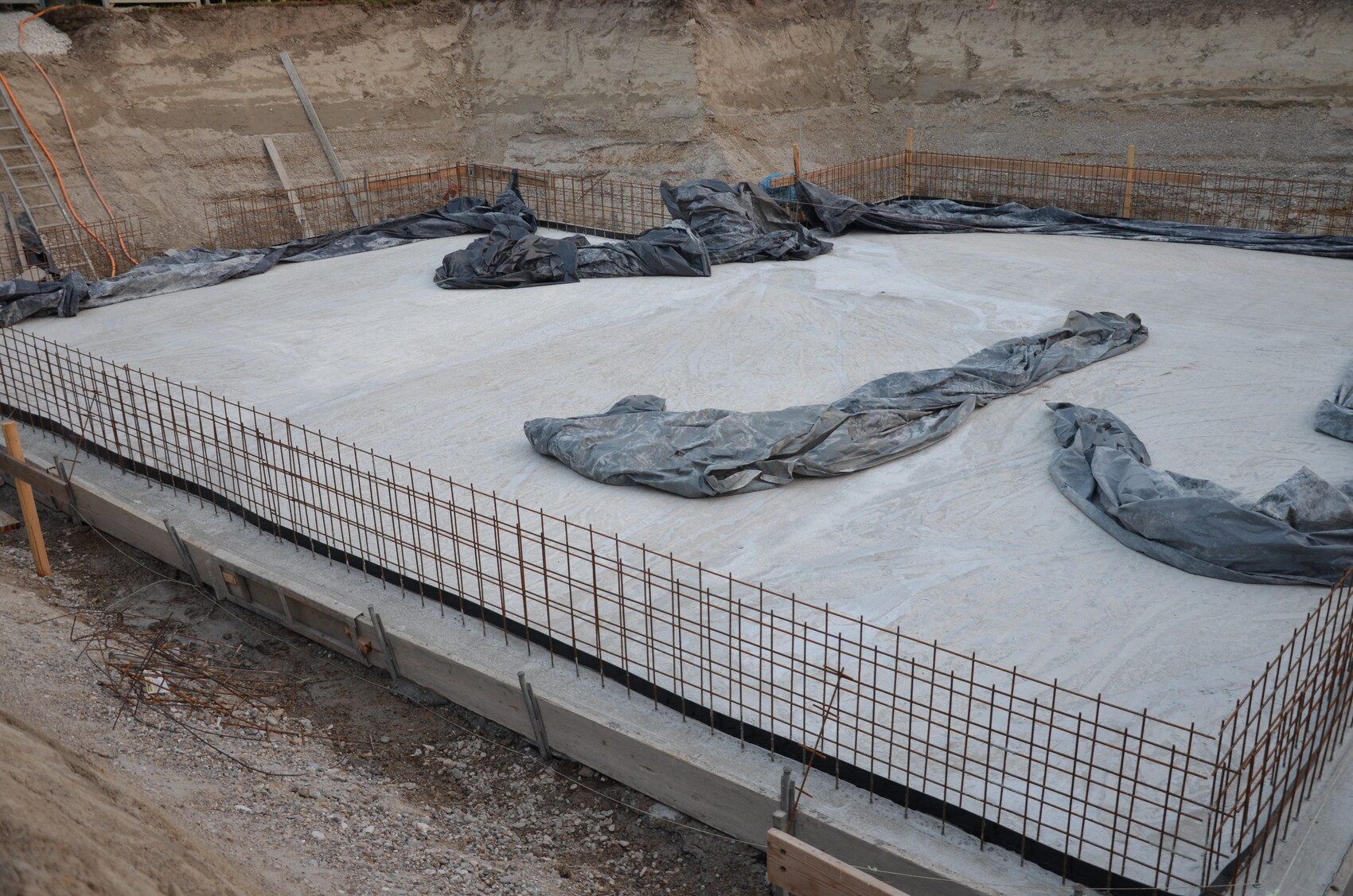 Baustelle: Betonfundament mit Bewehrungsstahlgitter, Abdeckplanen und Erdwand im Hintergrund.