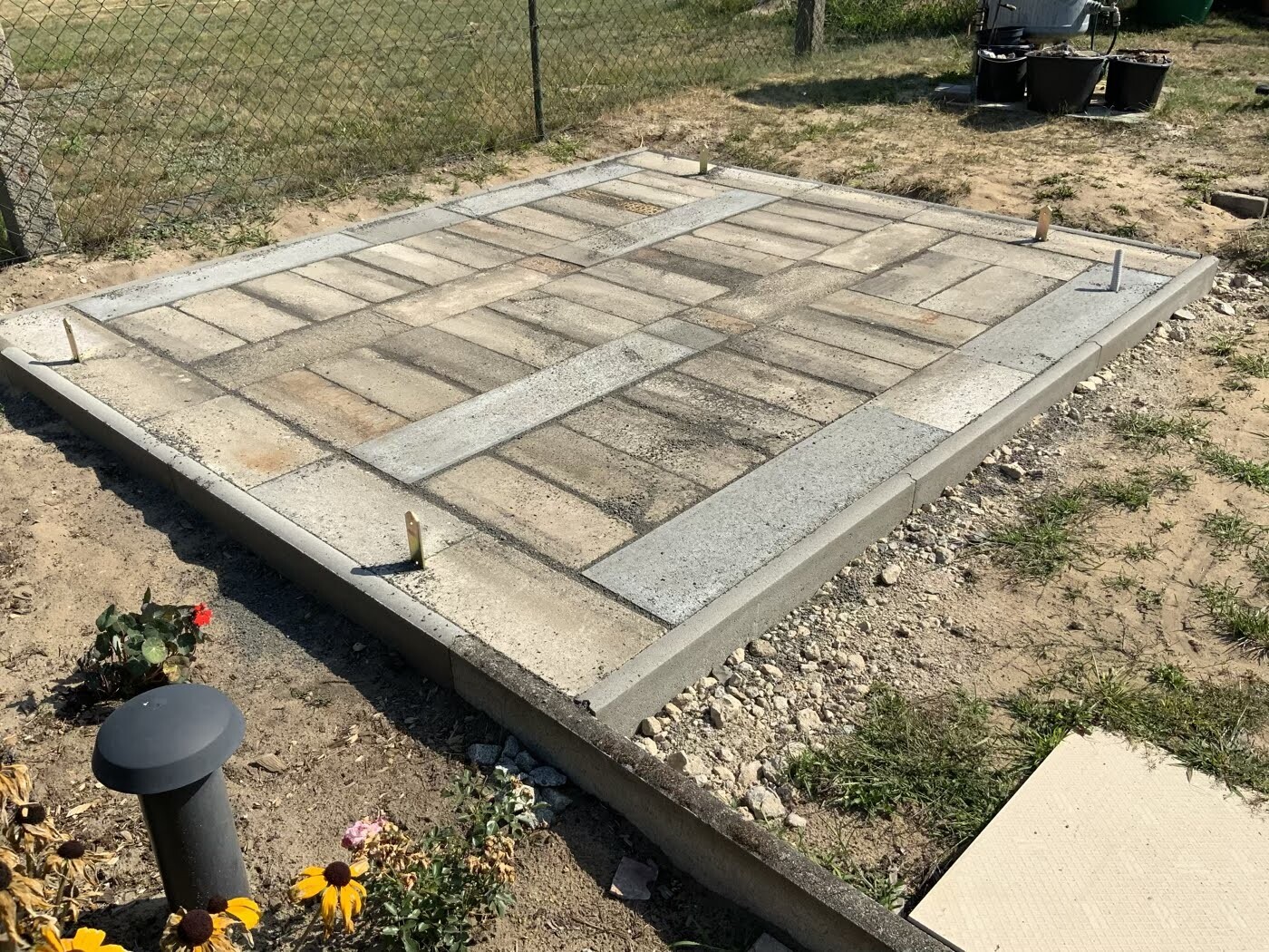 Betonplatte mit Pflastersteinen und Randsteinen; Garten mit Zaun und Blumen im Vordergrund.