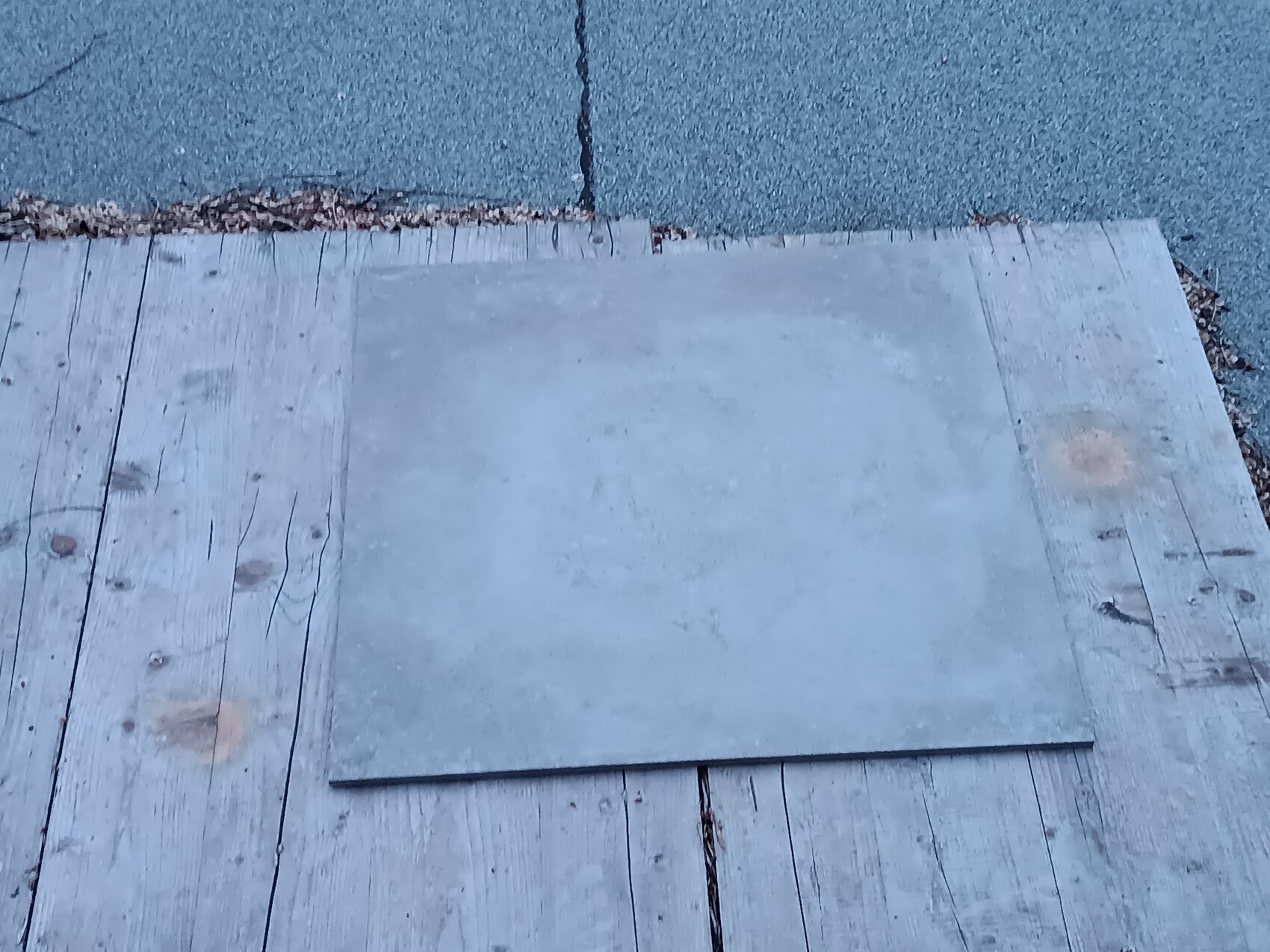 Quadratische Metallplatte liegt auf einem Holzdeck neben Asphaltweg.