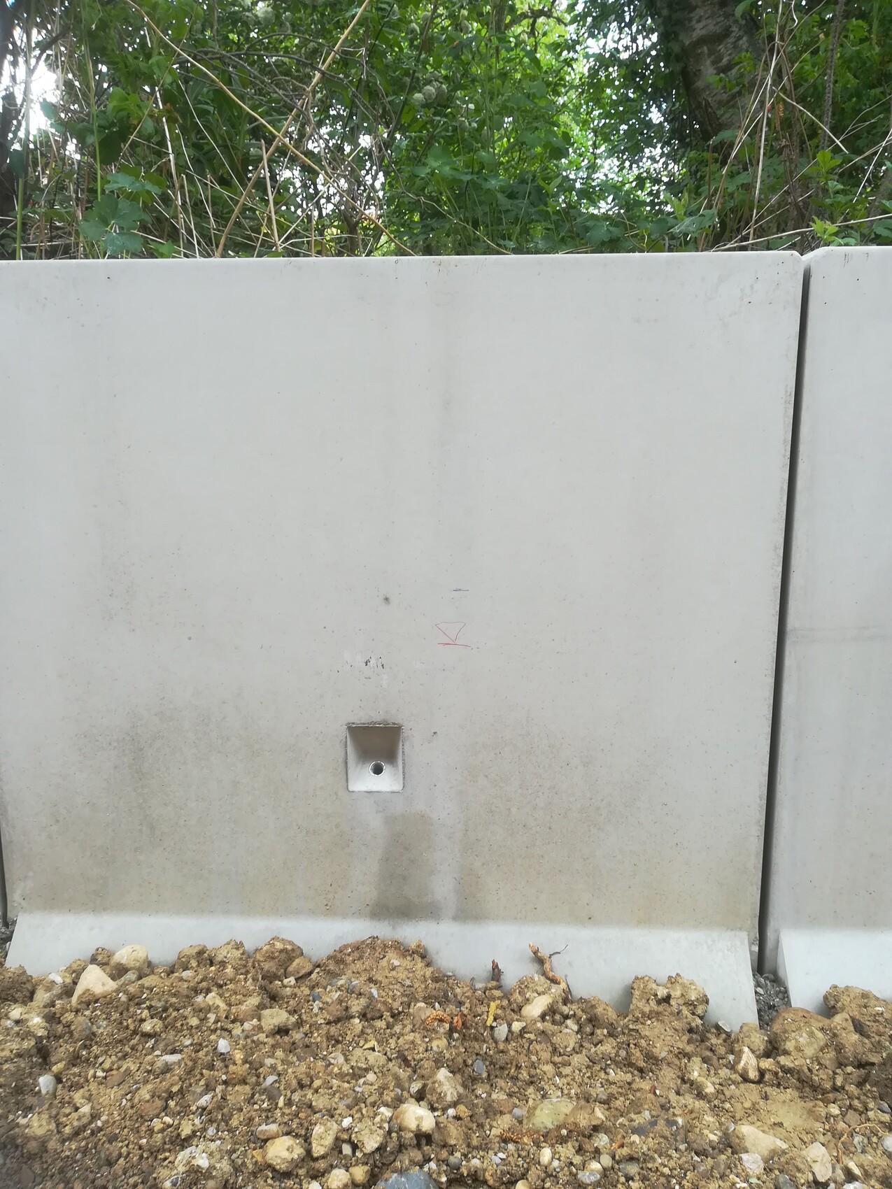Graue Betonbarriere mit quadratischer Metallbefestigung, Kies, Erdreich davor und grünem Hintergrund