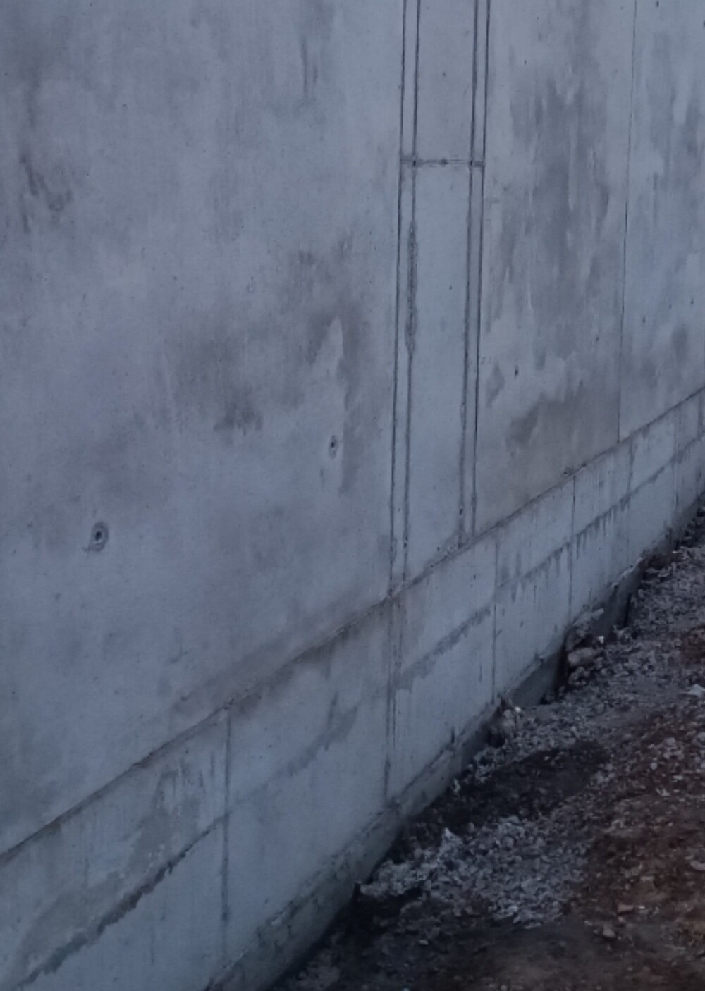 Graue Betonwand mit vertikalen Fugen, erdiger Untergrund und Kies am Boden.