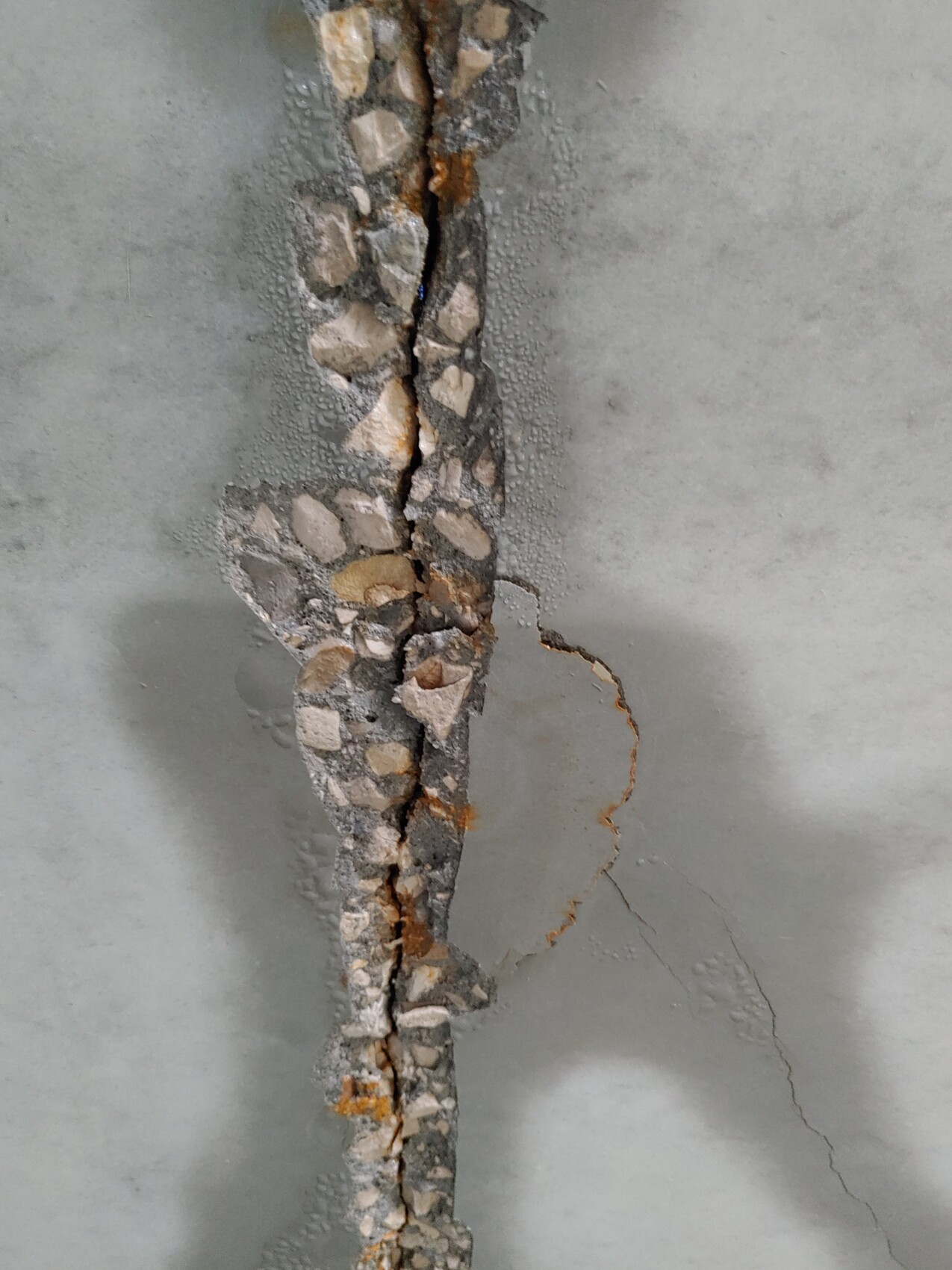 Vertikale Risse in einer Betonwand, freiliegte Kieselsteine und Rost an der Bruchstelle.