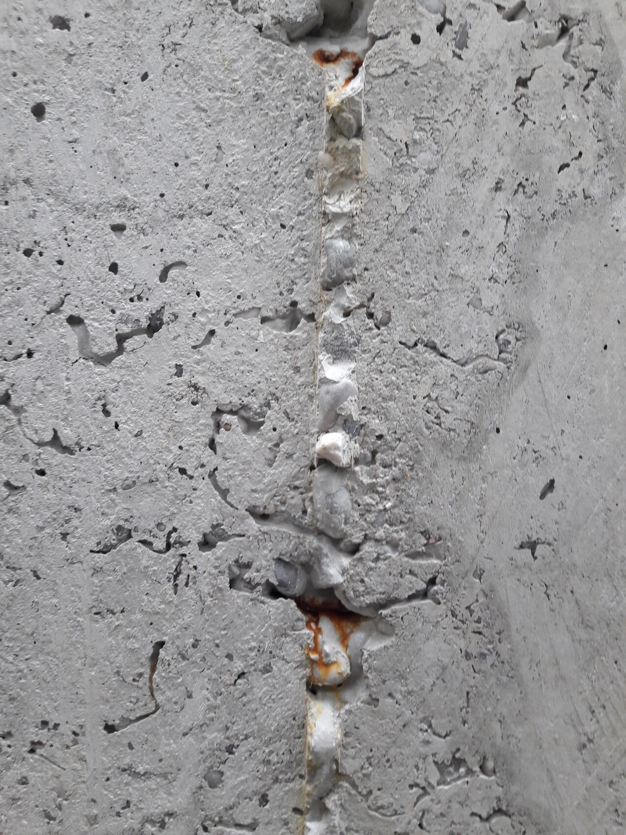 Rissige Betonwand mit vertikaler Fuge, Rostflecken und rauer Oberflächenstruktur.