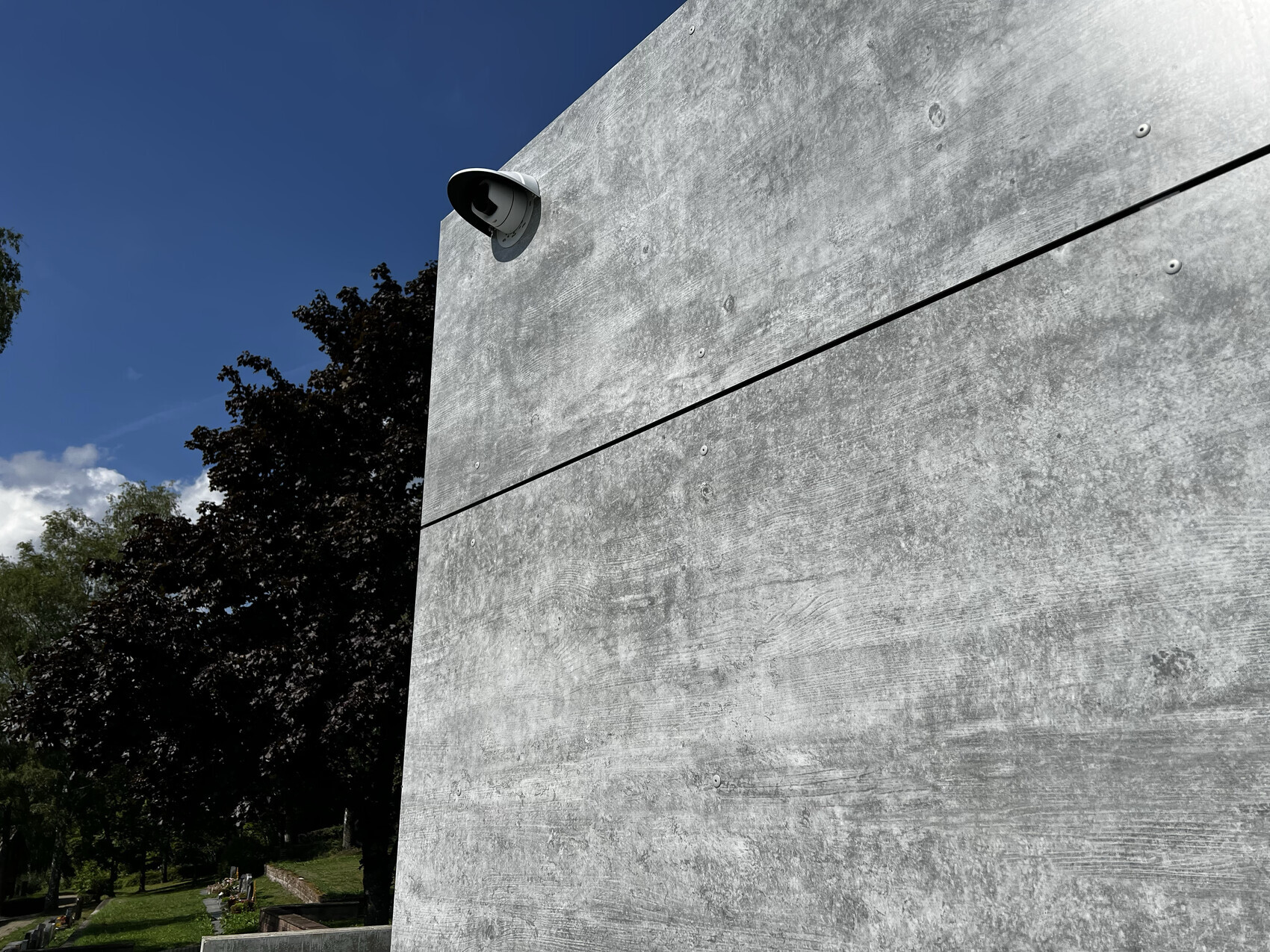 Große graue Betonwand mit Überwachungskamera oben links, blauer Himmel und Bäume im Hintergrund.