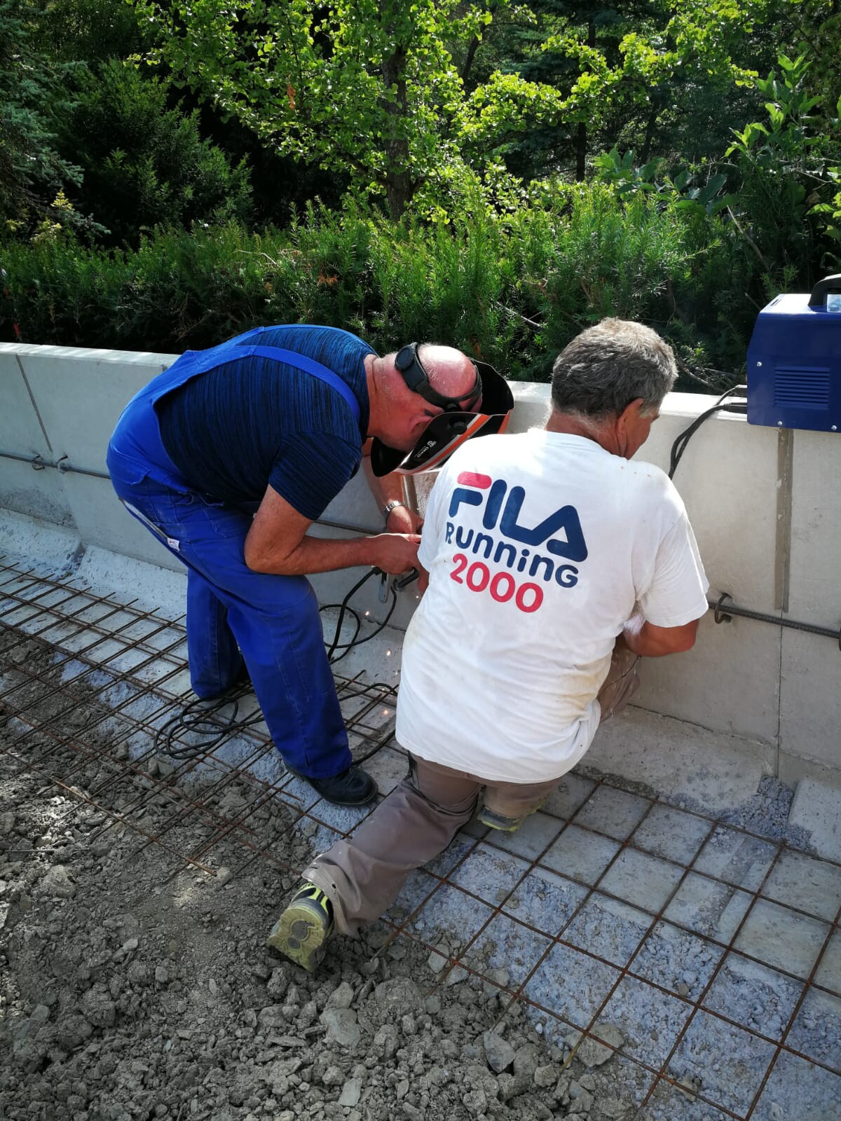 Zwei Bauarbeiter legen Stahlarmierung in eine Betonschalung im Freien, einer mit Visier.