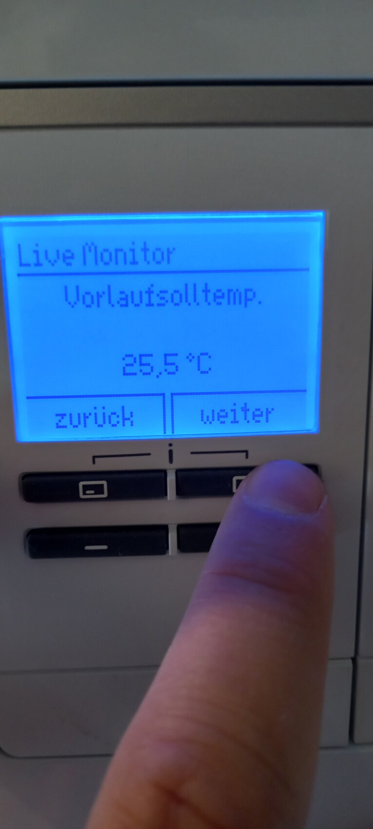 Blaues Display eines Thermostats mit Live Monitor, Vorlaufsolltemp. 25,5°C; Finger drückt 'weiter'.