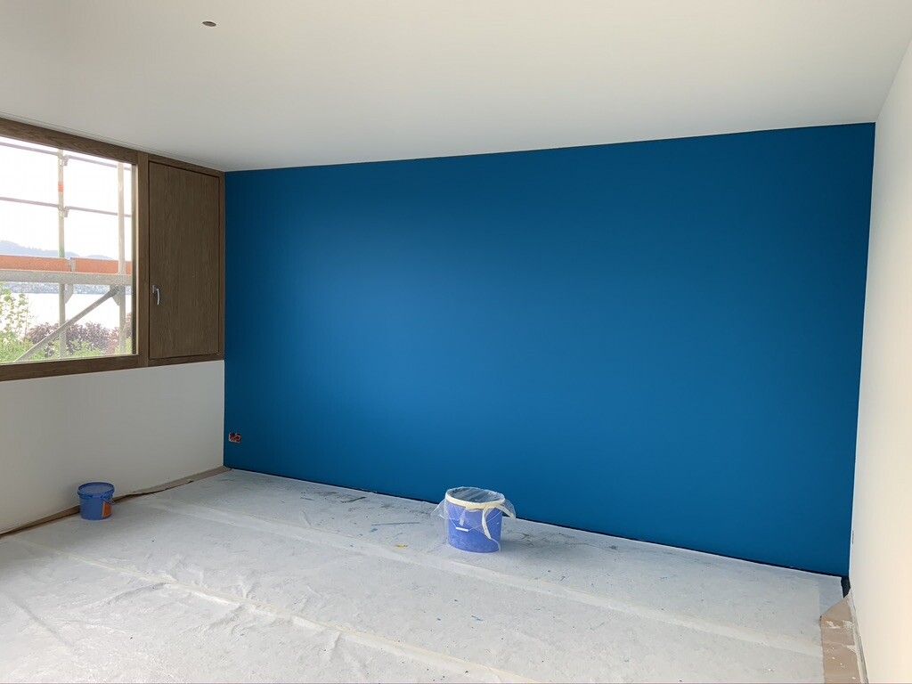 Raum mit blau gestrichener Akzentwand; Schutzfolie auf dem Boden, Eimer mit Farbe, Fenster links.