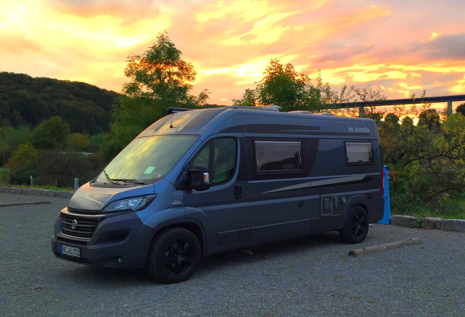 Hellblaues Wohnmobil auf Parkplatz, Abendhimmel in Orange, grüne Bäume dahinter.