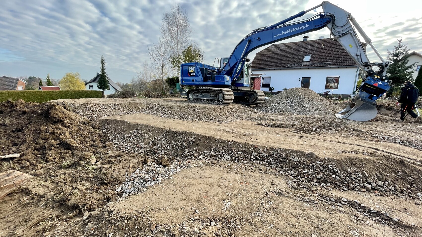 Großer blauer Kettenbagger arbeitet auf einer Baugrube mit Erd- und Kieshügeln, Wohnhaus dahinter.