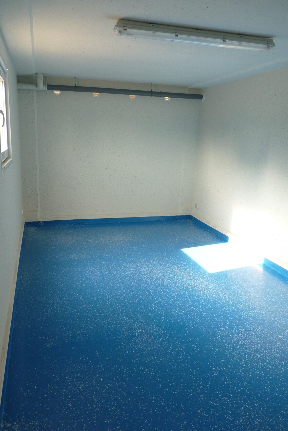 Leerer Raum mit blau gesprenkeltem Boden, weißen Wänden, Deckenleuchte und Fenster.