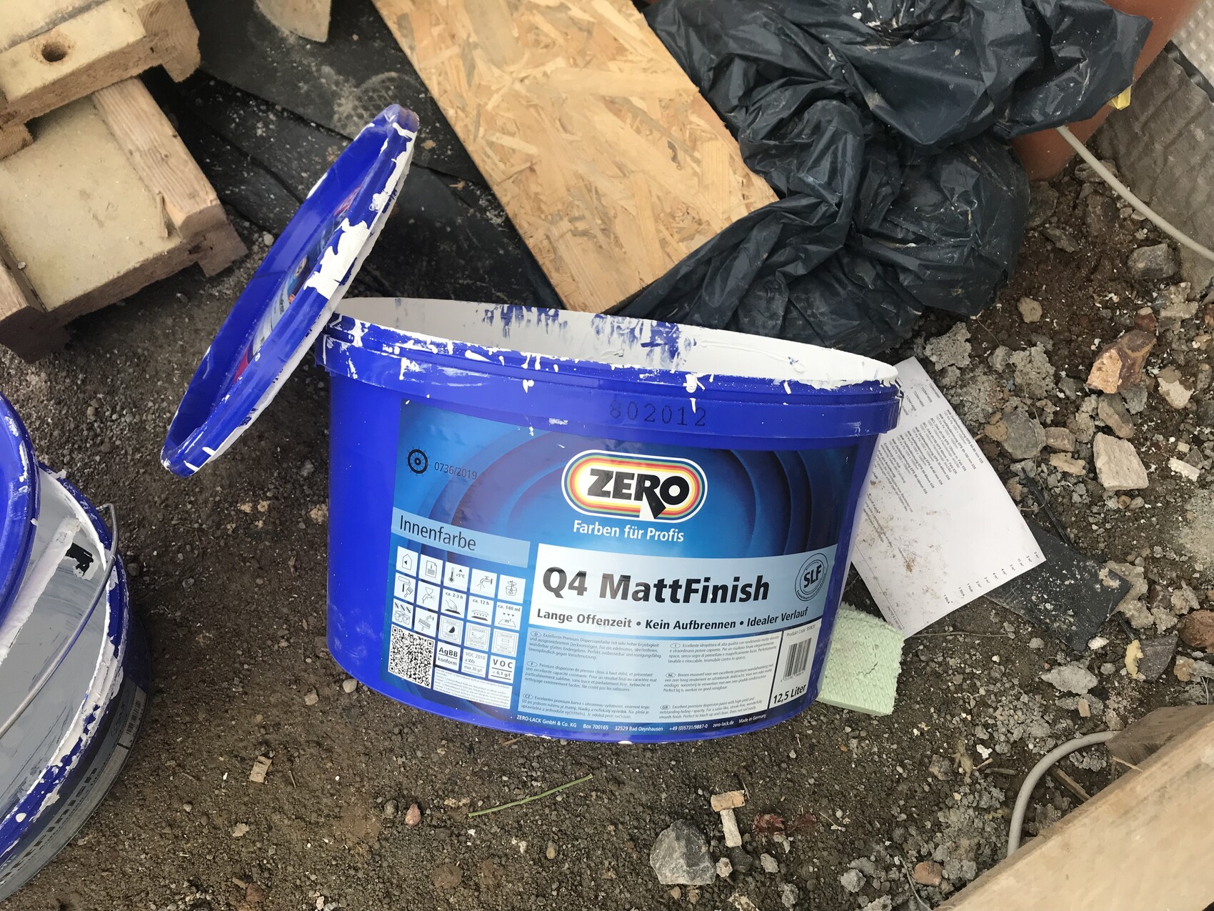 Offener blauer Farbeimer Q4 MattFinish auf Baustelle, daneben Holzreste und Müll.