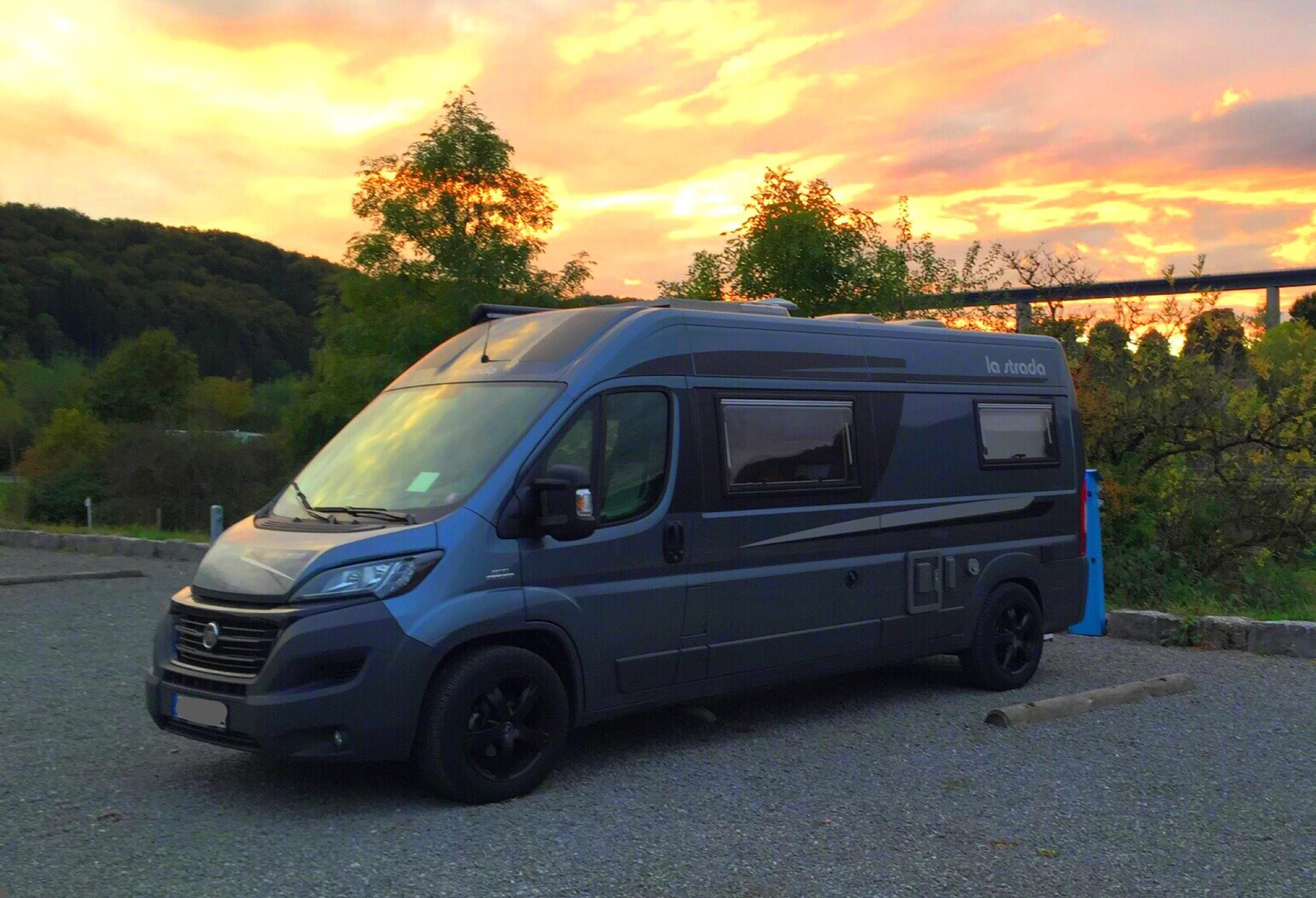 Blaues La Strada Wohnmobil parkt auf einem Kiesparkplatz bei Sonnenuntergang, Bäume im Hintergrund.
