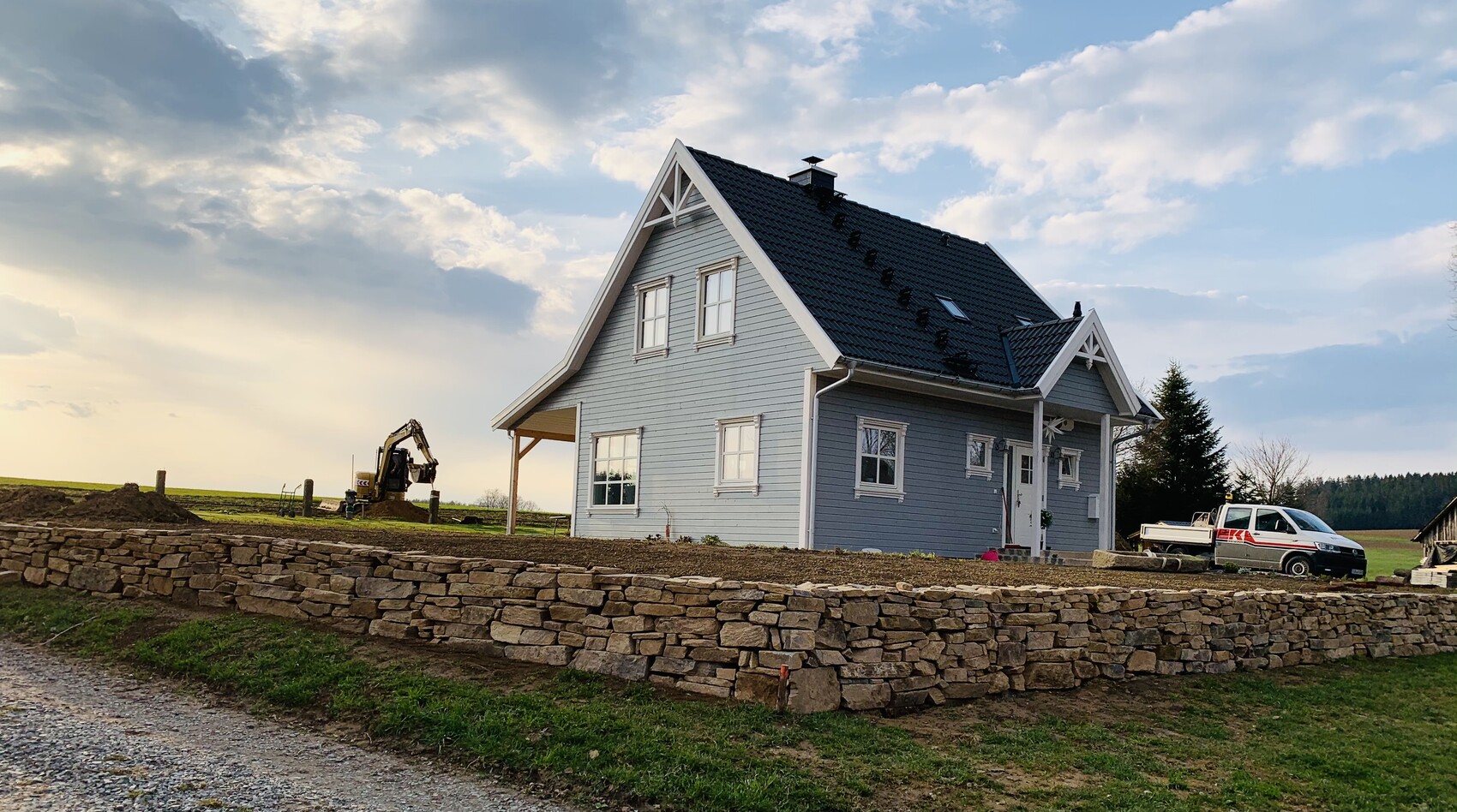 Graues Einfamilienhaus mit dunklem Dach hinter Steinmauer; Baugerät und Transporter im Feld.
