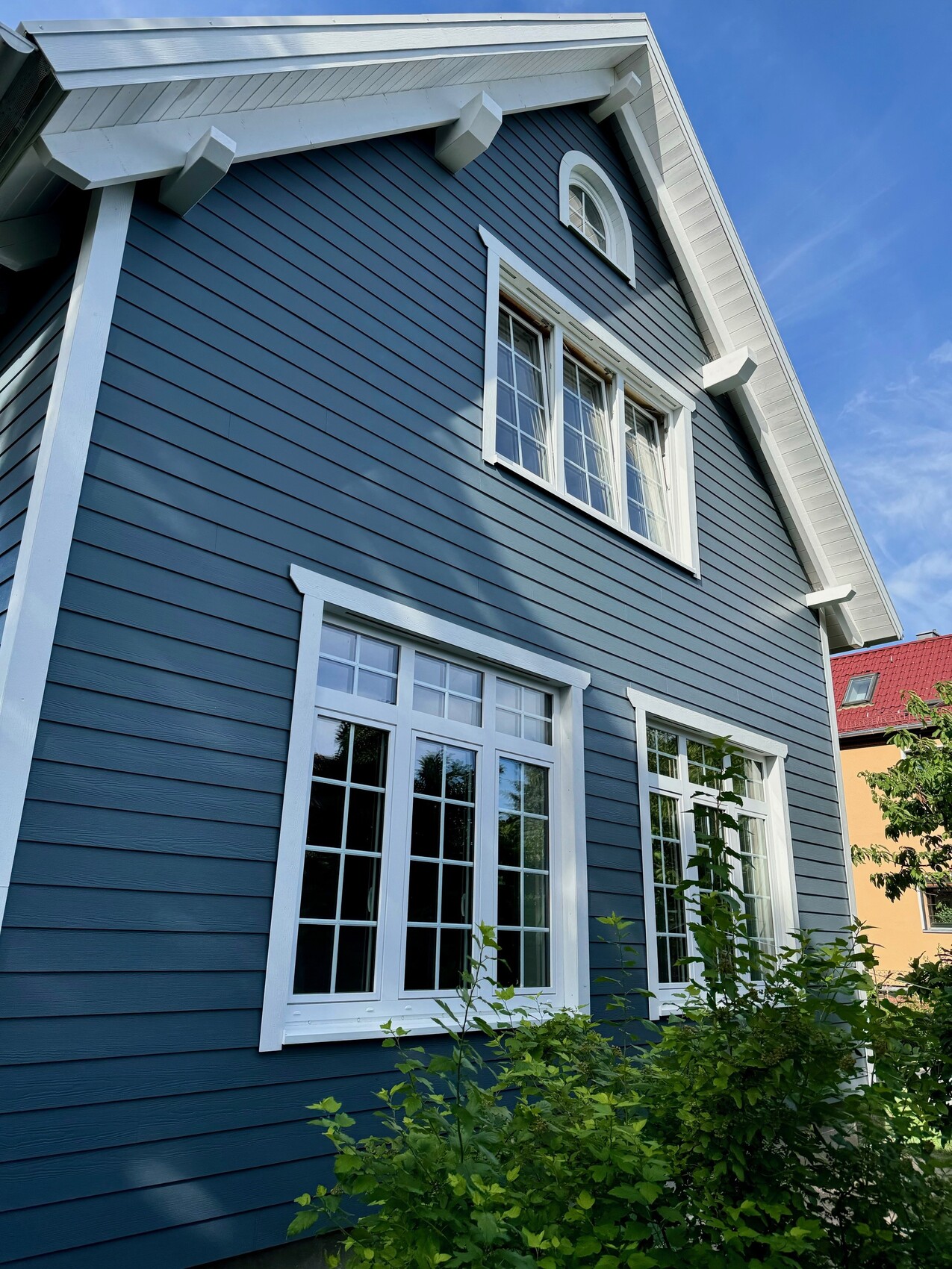Zweistöckiges blaues Holzhaus mit weißen Fensterrahmen und grünem Sträuchern davor.