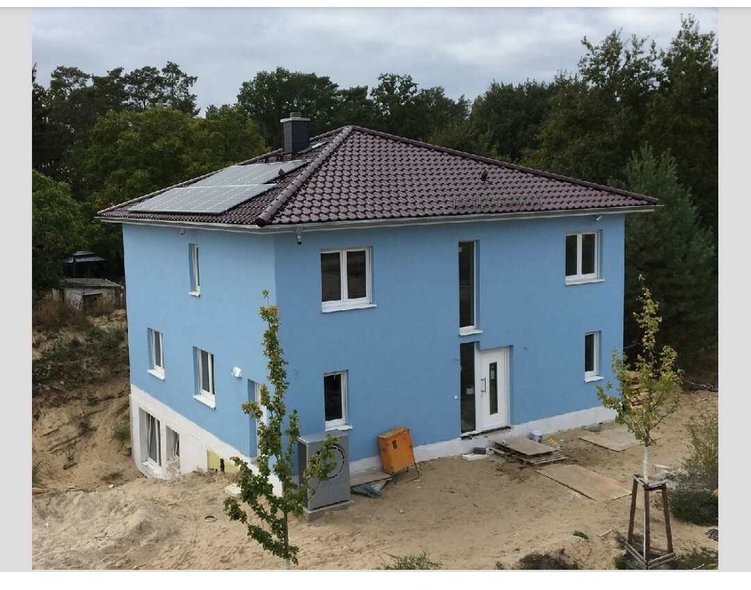 Zweistöckiges blaues Haus mit Solarpanelen auf dem Dach; Baugrund umgeben von Bäumen.