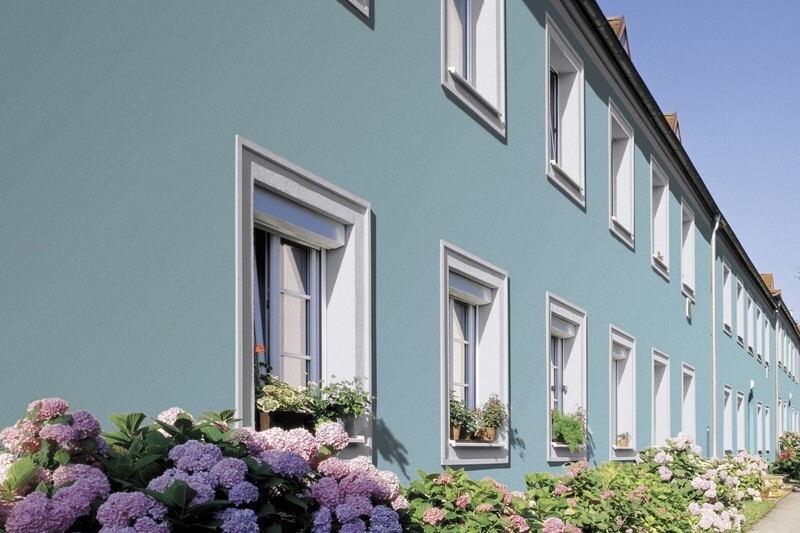 Blaue Fassade eines Hauses mit weißen Fensterrahmen und blühenden Blumen im Vorgarten