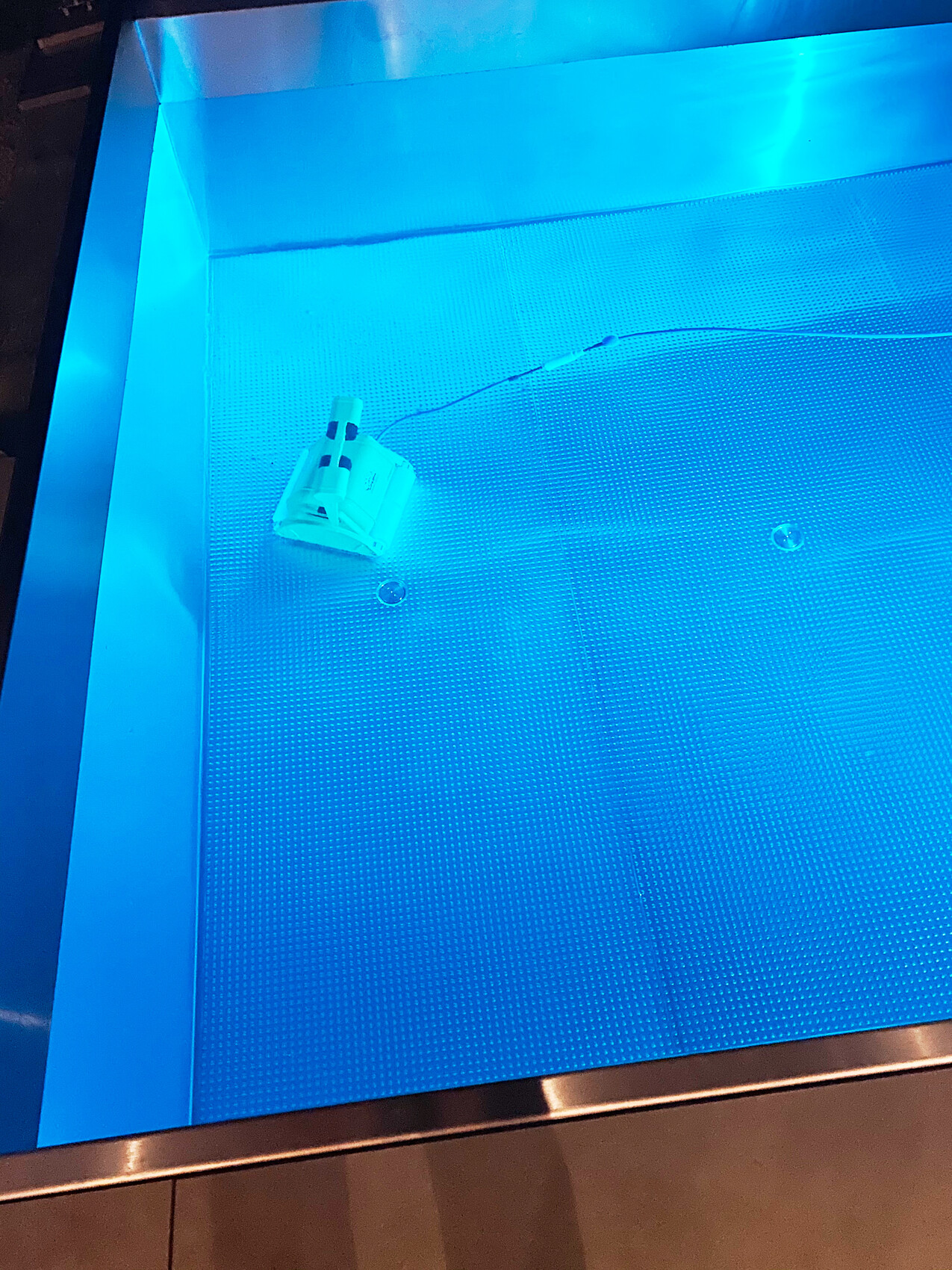 Blau beleuchteter Wassertank; am Boden liegt ein weißes, kabelgebundenes Elektronikmodul.