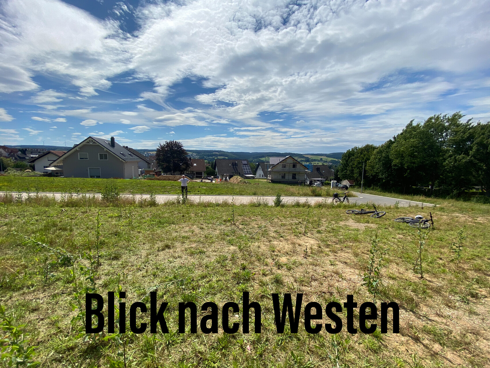 Feld mit Häusern am Horizont, zwei Fahrräder liegen, Person am Straßenrand, blauer Himmel.