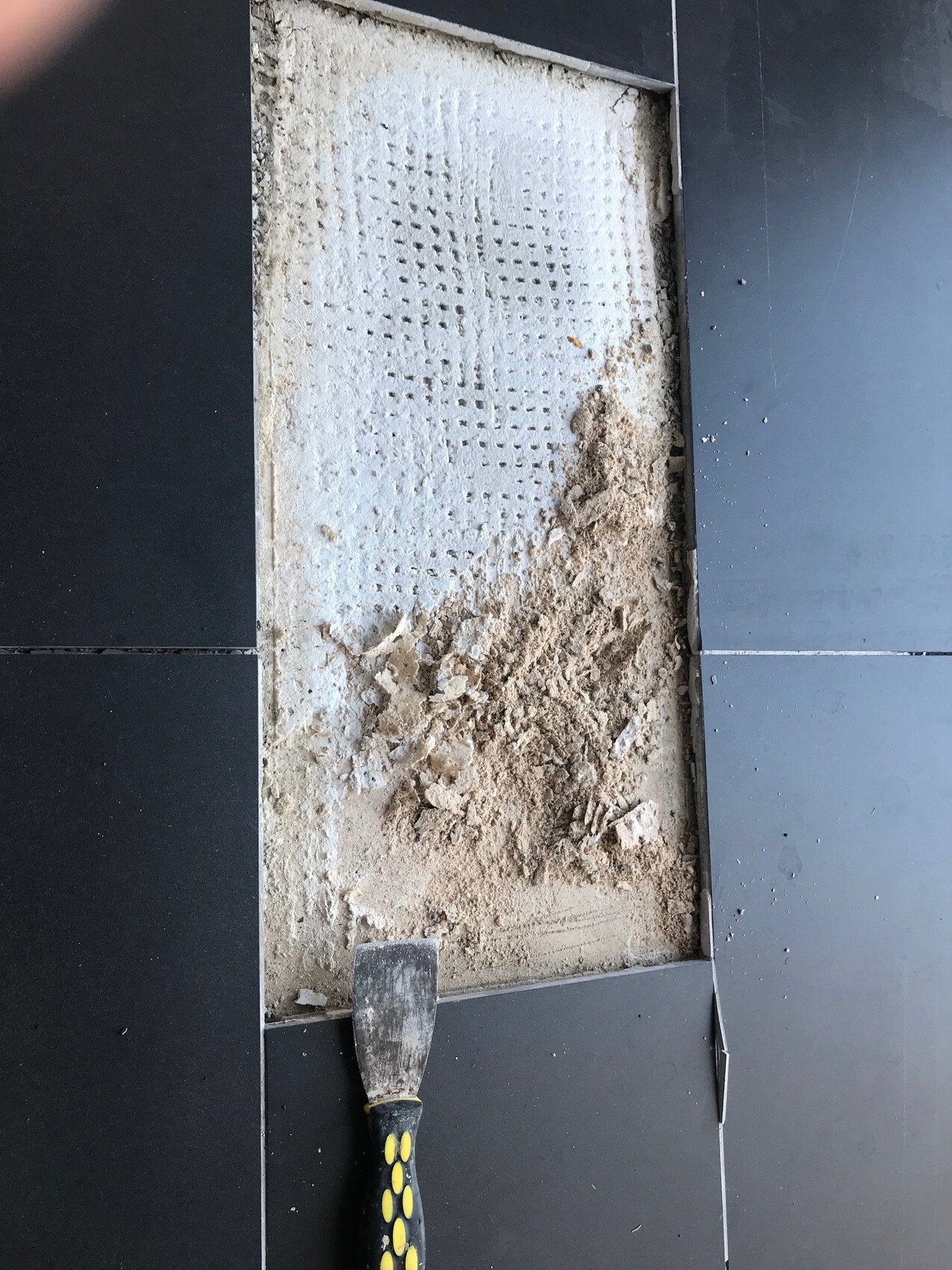 Dunkler Fliesenboden mit Aussparung, Sand und weiße perforierte Unterlage; Spachtel am Rand.