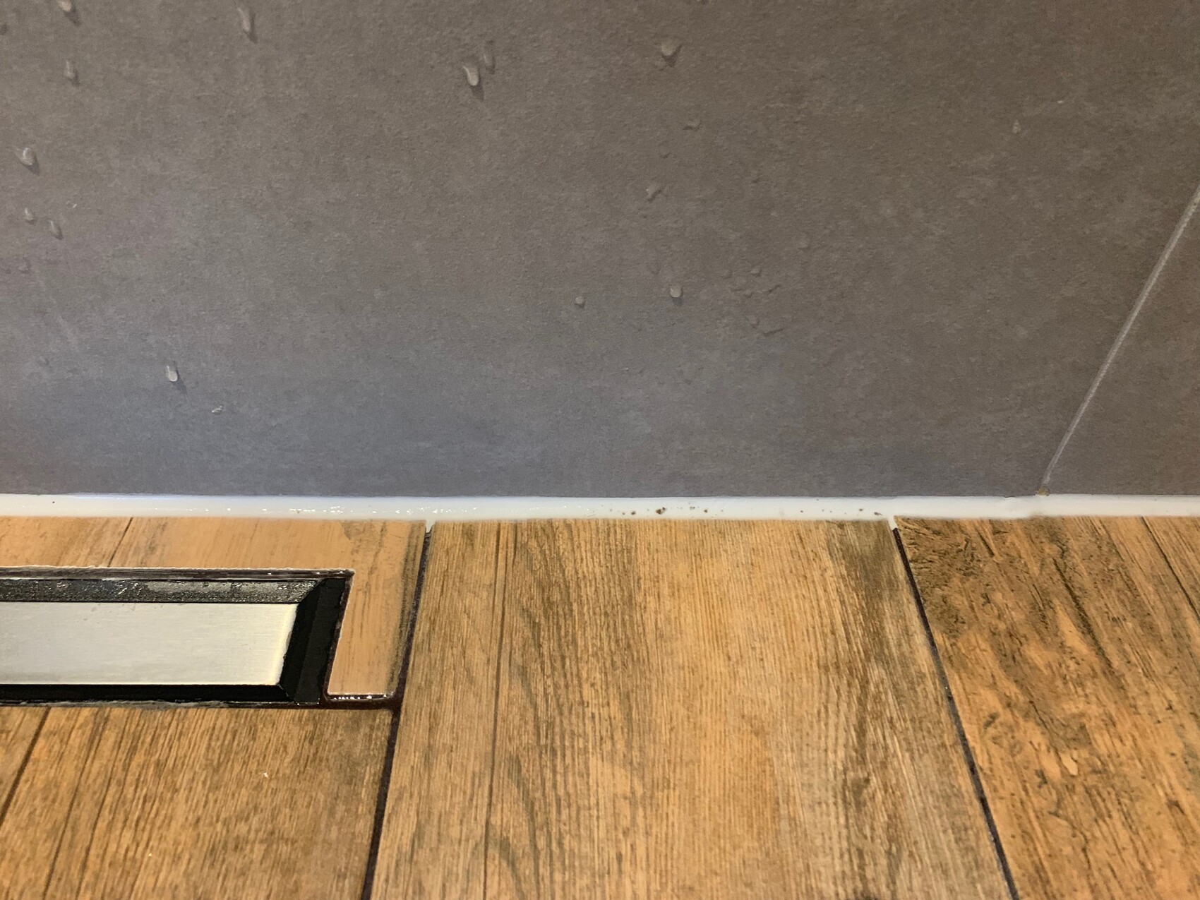 Nahaufnahme eines Bodens mit Holzoptik-Fliesen und Fußleiste an grauer Wand.
