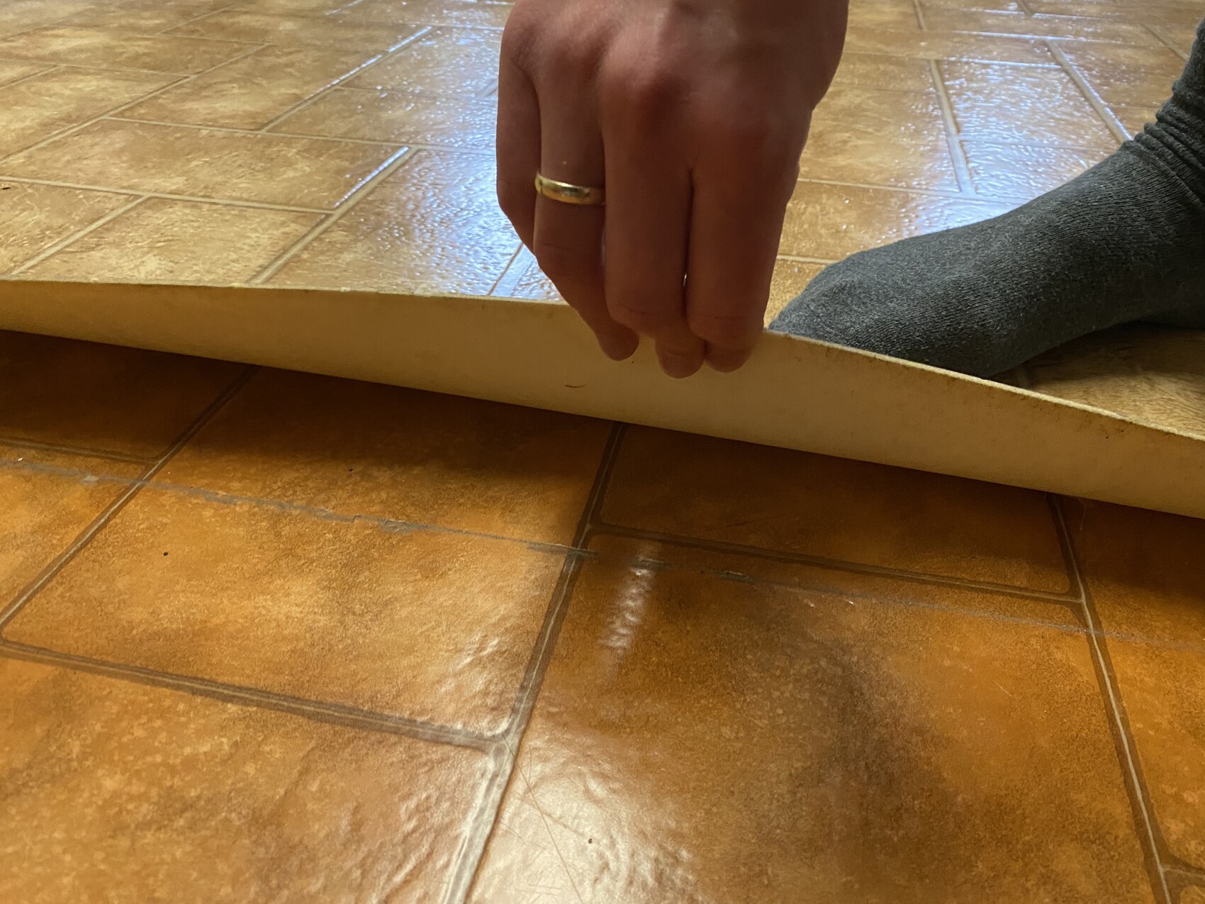 Hand mit Ring bewegt eine dünne Holzleiste über braune Fliesen; graue Socke am Fuß sichtbar.