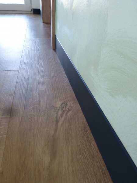 Nahaufnahme eines Laminatbodens im Flur neben grüner Wand und dunkler Sockelleiste.
