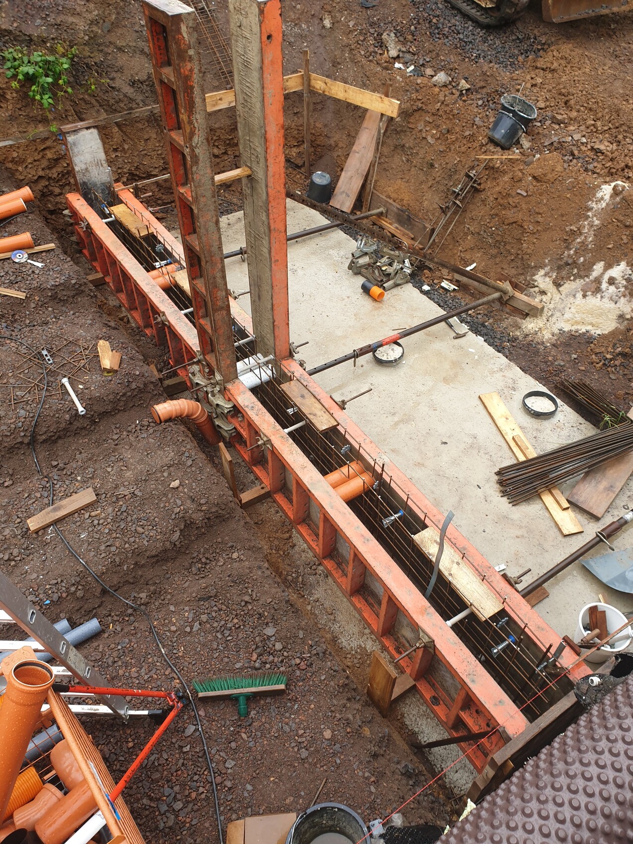 Baustelle mit roter Stahl-Schalung, Bewehrung und Holzrahmen um eine Betonfundamentplatte im Erdreich.