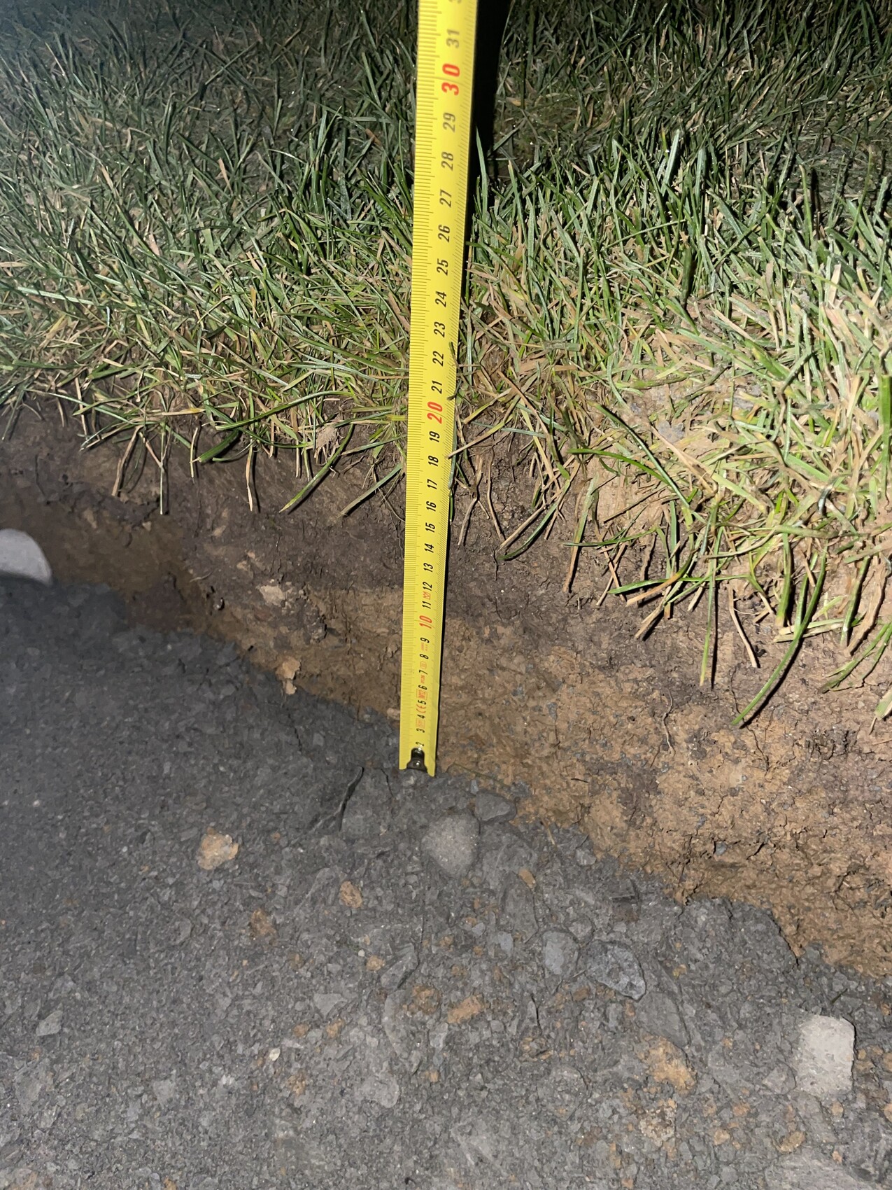 Messband misst Baugrundtiefe neben Grasrand und Asphaltboden.