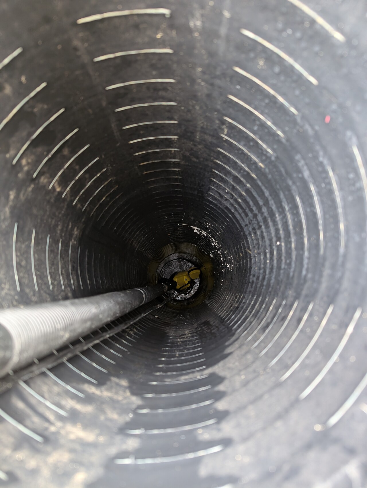 Tiefer, dunkler Schacht mit spiralförmiger Innenwand; Schlauch liegt drin, gelbes Werkzeug am Boden.