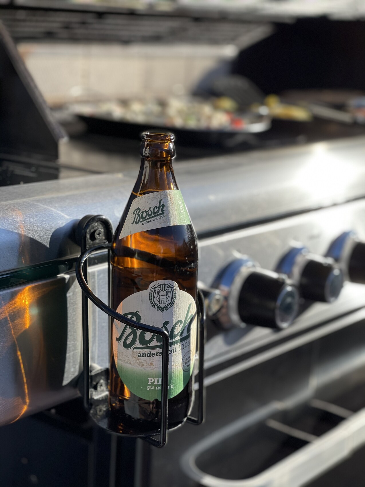Eine braune Bierflasche mit Bosch-Etikett hängt in einem Metallhalter am Grill.