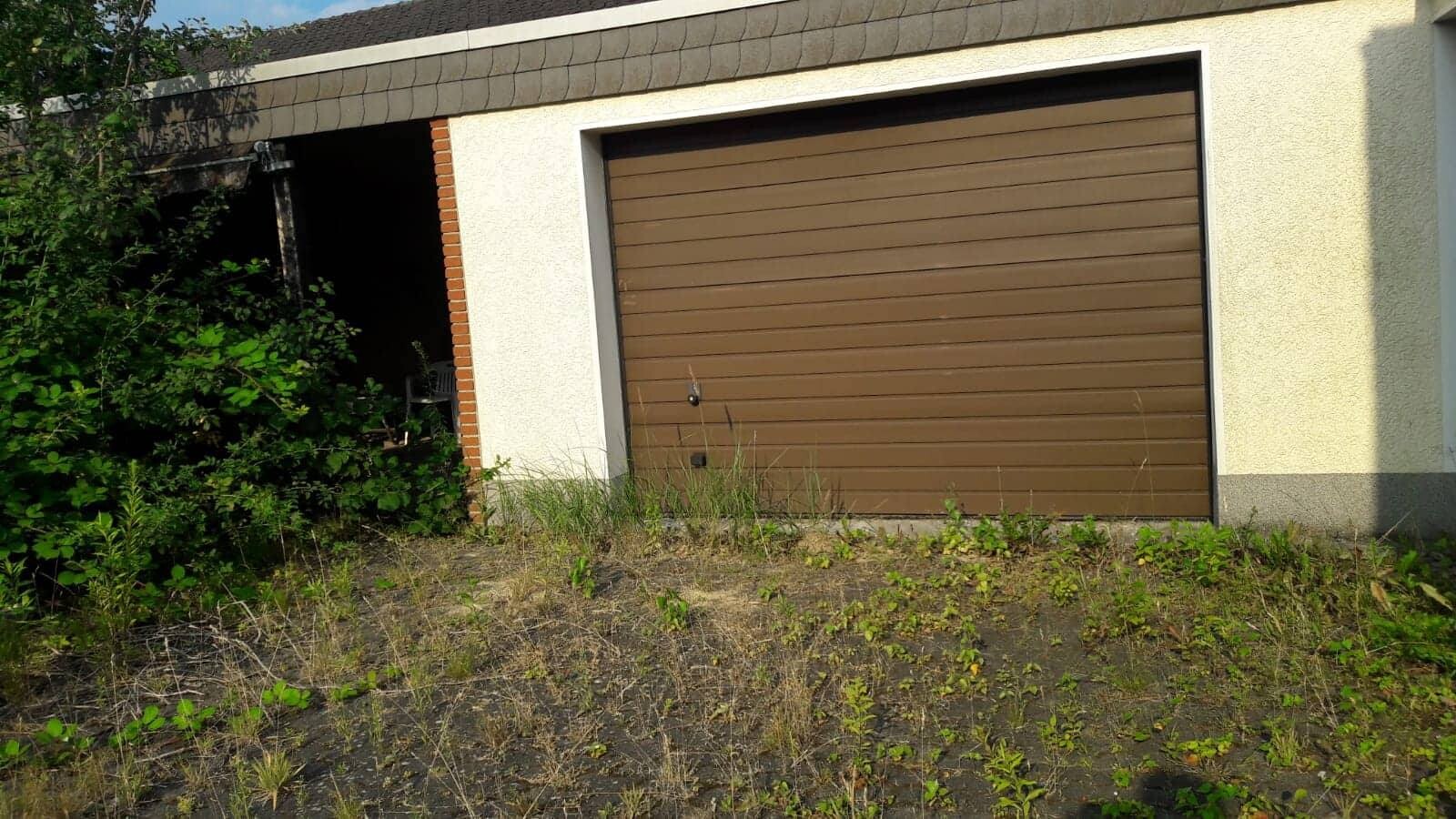 Braune Garagenrolltor an einer hellen Fassade; links dunkler Durchgang, davor Unkraut und Gras.