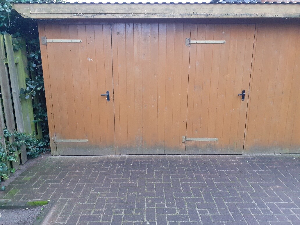 Großer brauner Holzschuppen mit zwei Türen und Metallbeschlägen vor gepflastertem Hof.