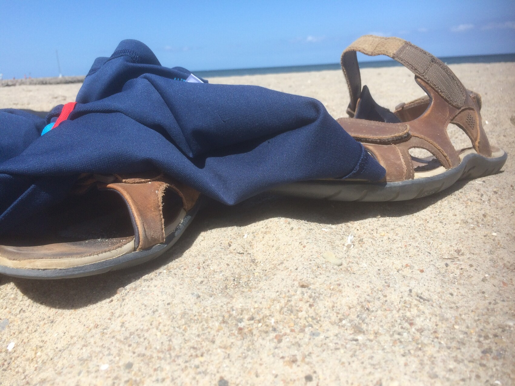 Braune Sandalen liegen im Sand am Strand, daneben blaues Kleidungsstueck, Blick aufs Meer.