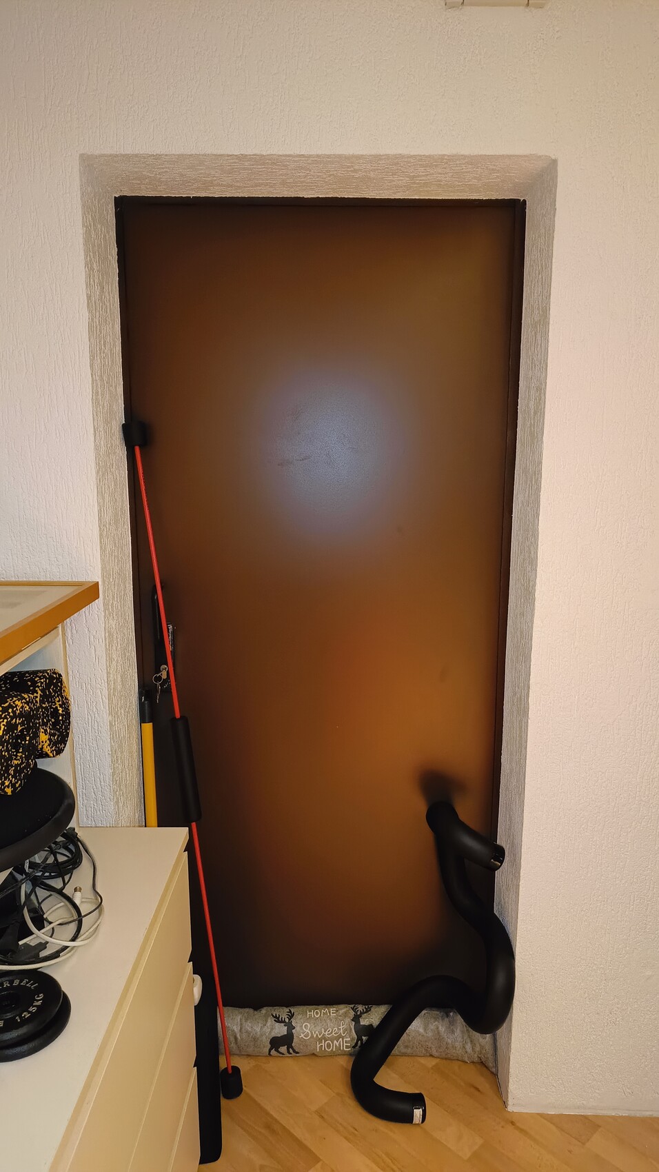Braune Tür in weißem Rahmen; rote Stange, schwarzer Schlauch, Kissen 'Home Sweet Home' am Boden.
