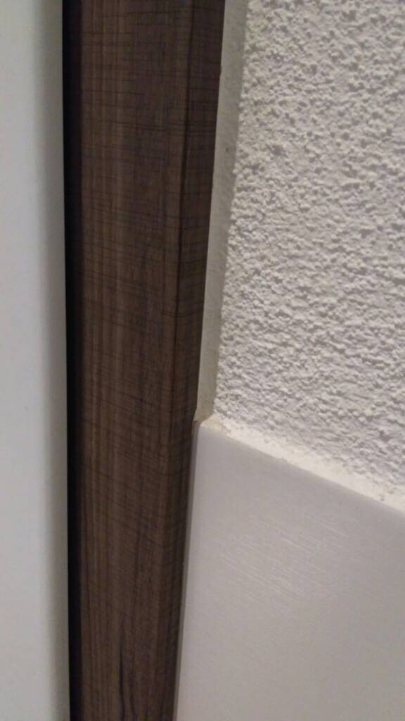 Nahaufnahme eines Türrahmens aus braunem Holzprofil neben einer weißen, rauen Wand