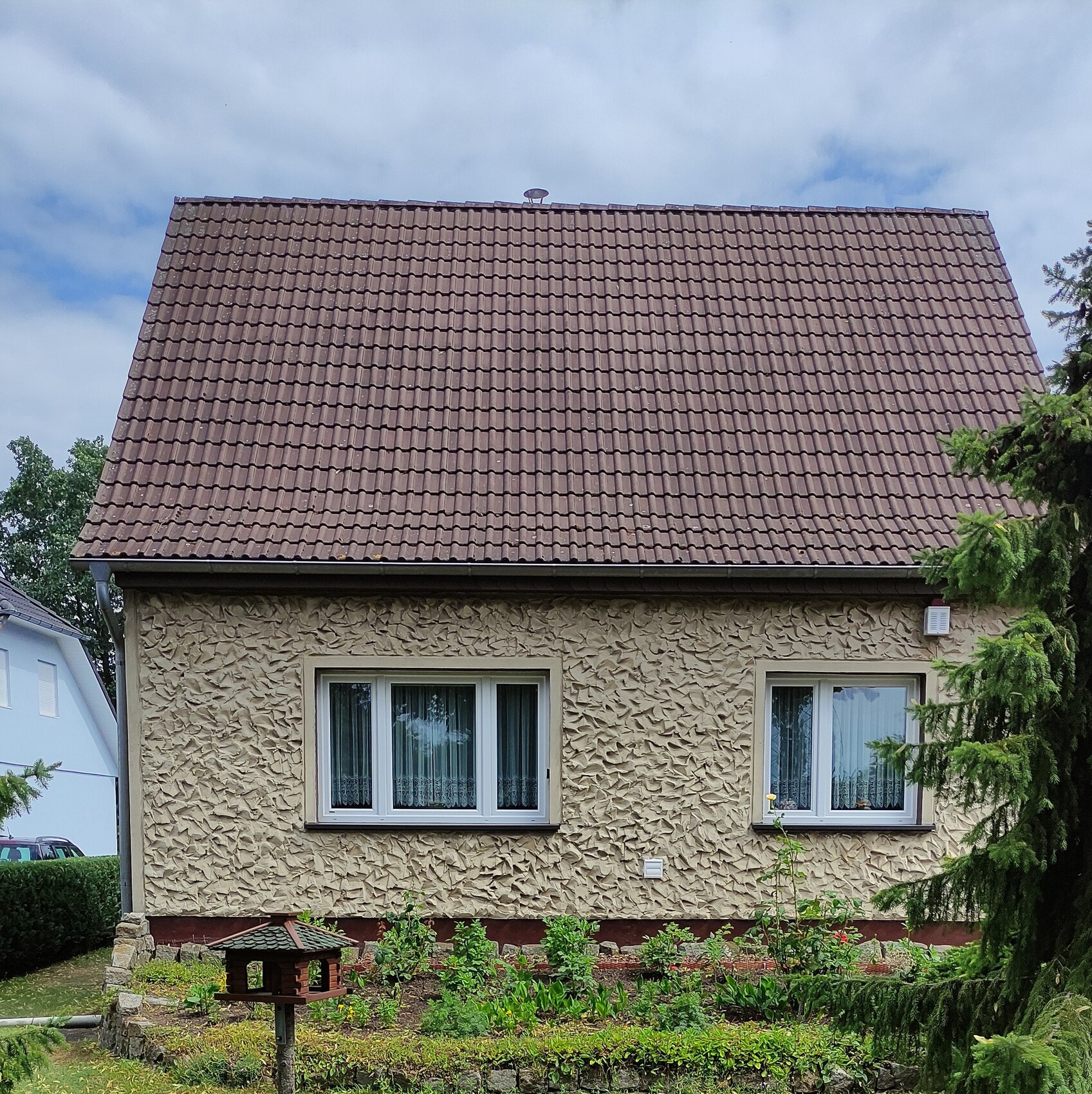 Häuschen mit braunem Satteldach, hell verputzter Fassade, zwei Fenster, Garten mit Vogelhaus.