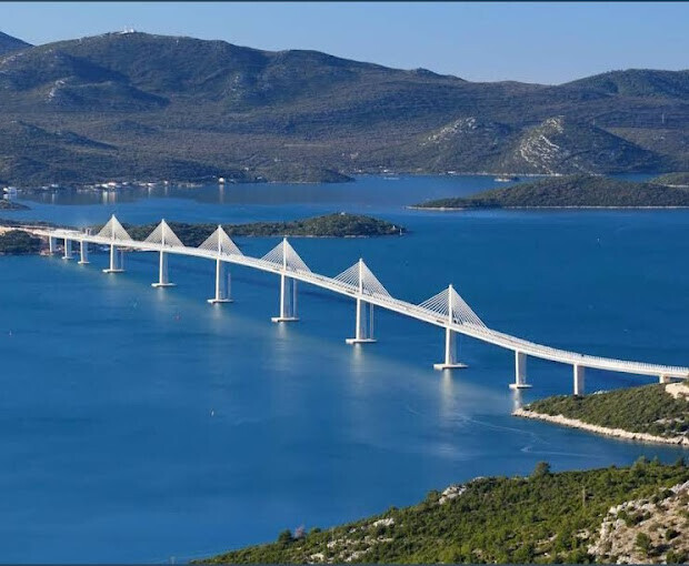 Weiße Seilbrücke mit Türmen zieht sich über blaues Meer, Berge im Hintergrund.