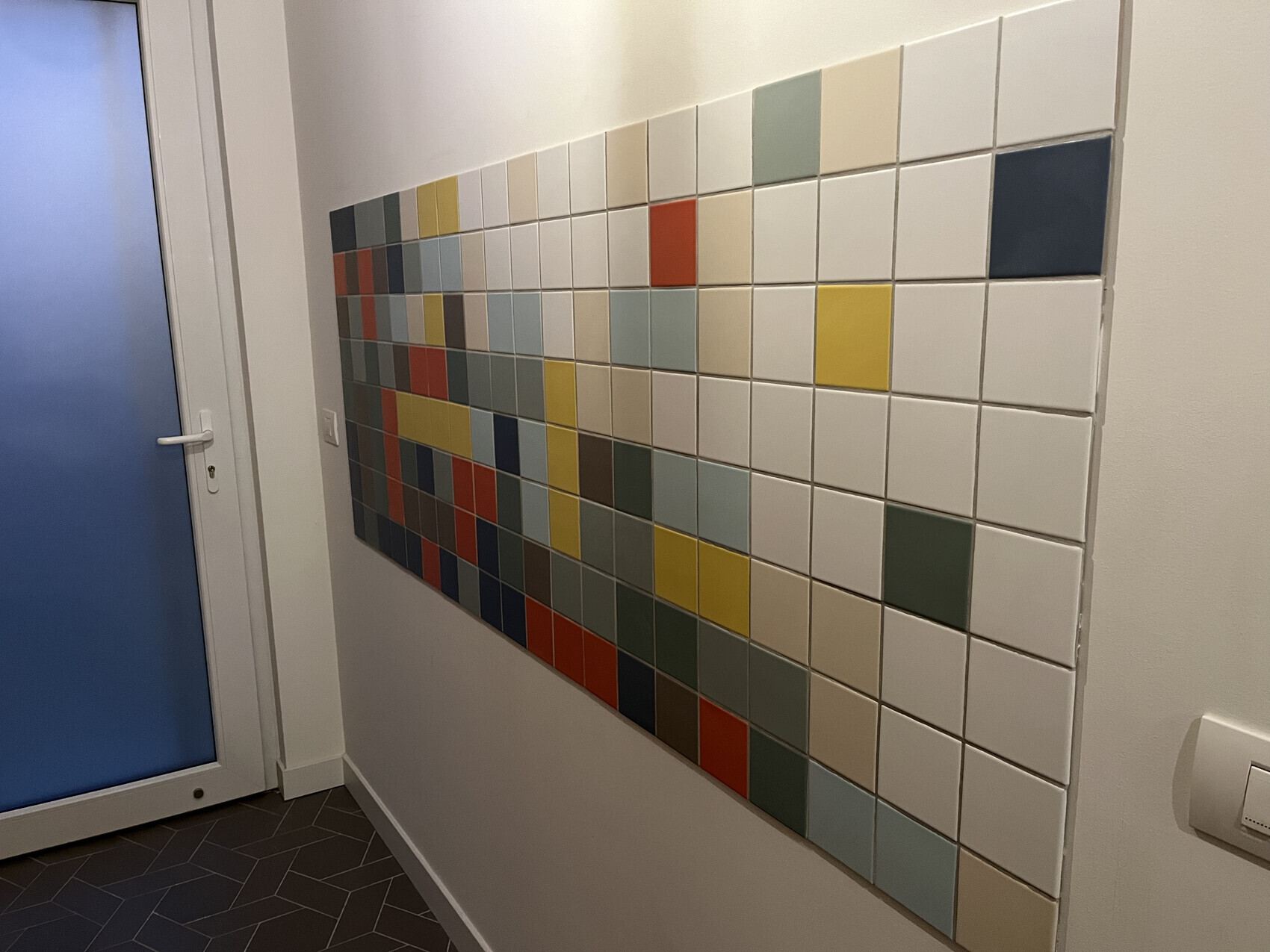 Wandpanel aus bunt gemusterten Quadratfliesen neben einer Tür mit blauem Glas.