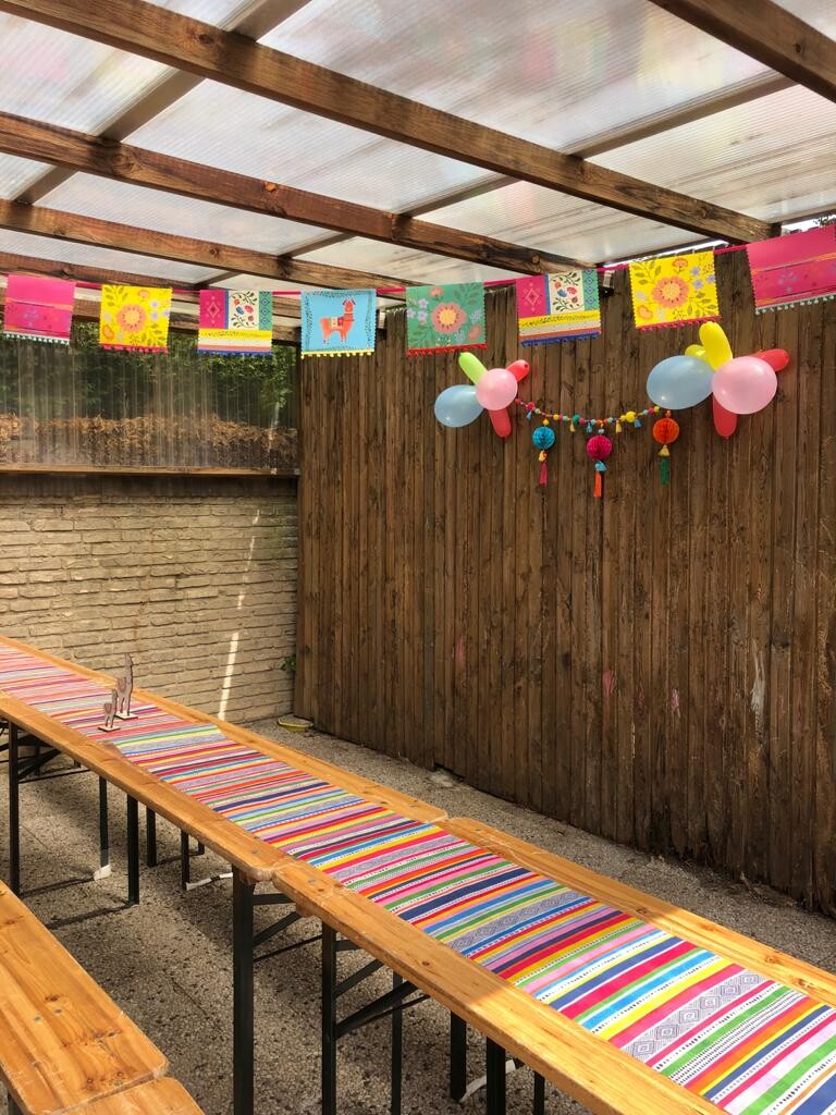 Lange Holztische mit bunten Tischläufern, bunte Banner und Ballons – Festdekoration im Freien.