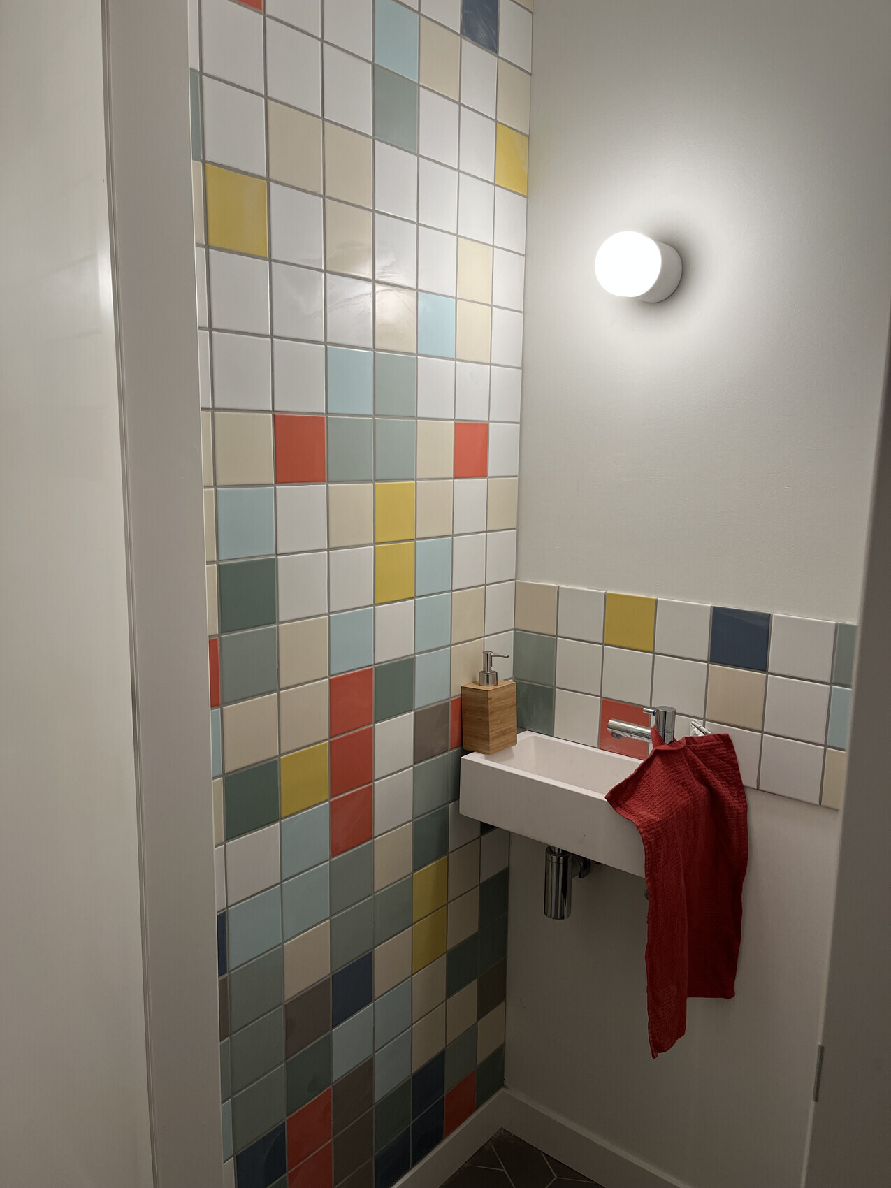 Kleines Waschbecken, bunte Pastellfliesen, rotes Handtuch, Holzseifenspender, runde Leuchte.