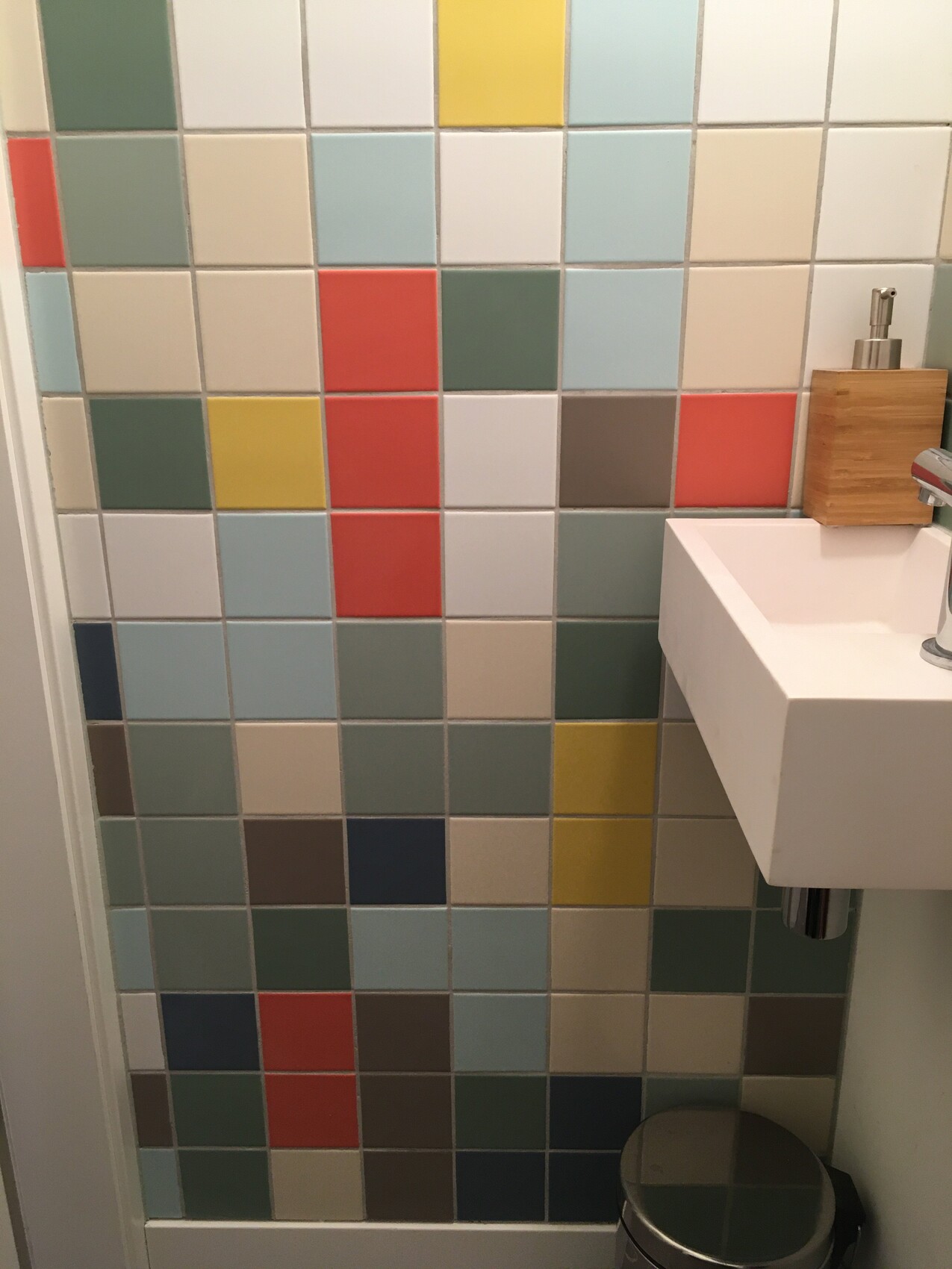 Bunte quadratische Fliesenwand im Badezimmer, weißes Waschbecken, Holz-Seifenspender, Müllbehälter.