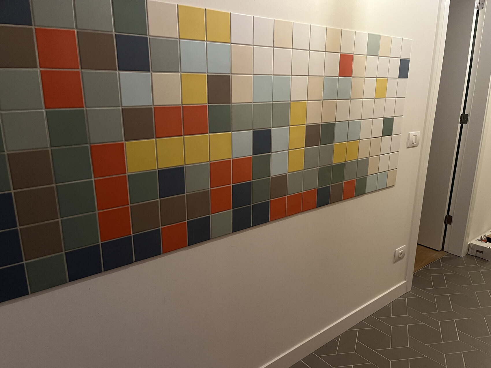 Großes quadratisches Fliesenmosaik in bunten Farben an einer Wand im Flur.