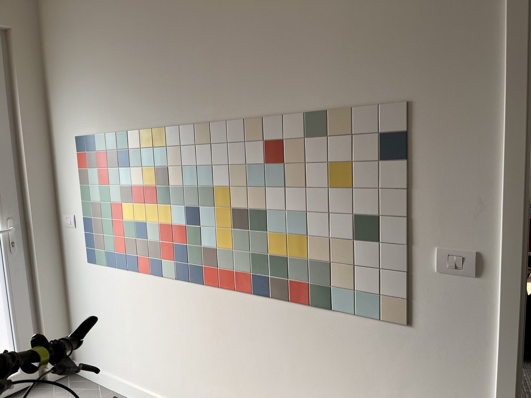 Großes farbiges Quadrat-Mosaik an weißer Wand, abstrakte Kunst; Fahrradteile sichtbar.