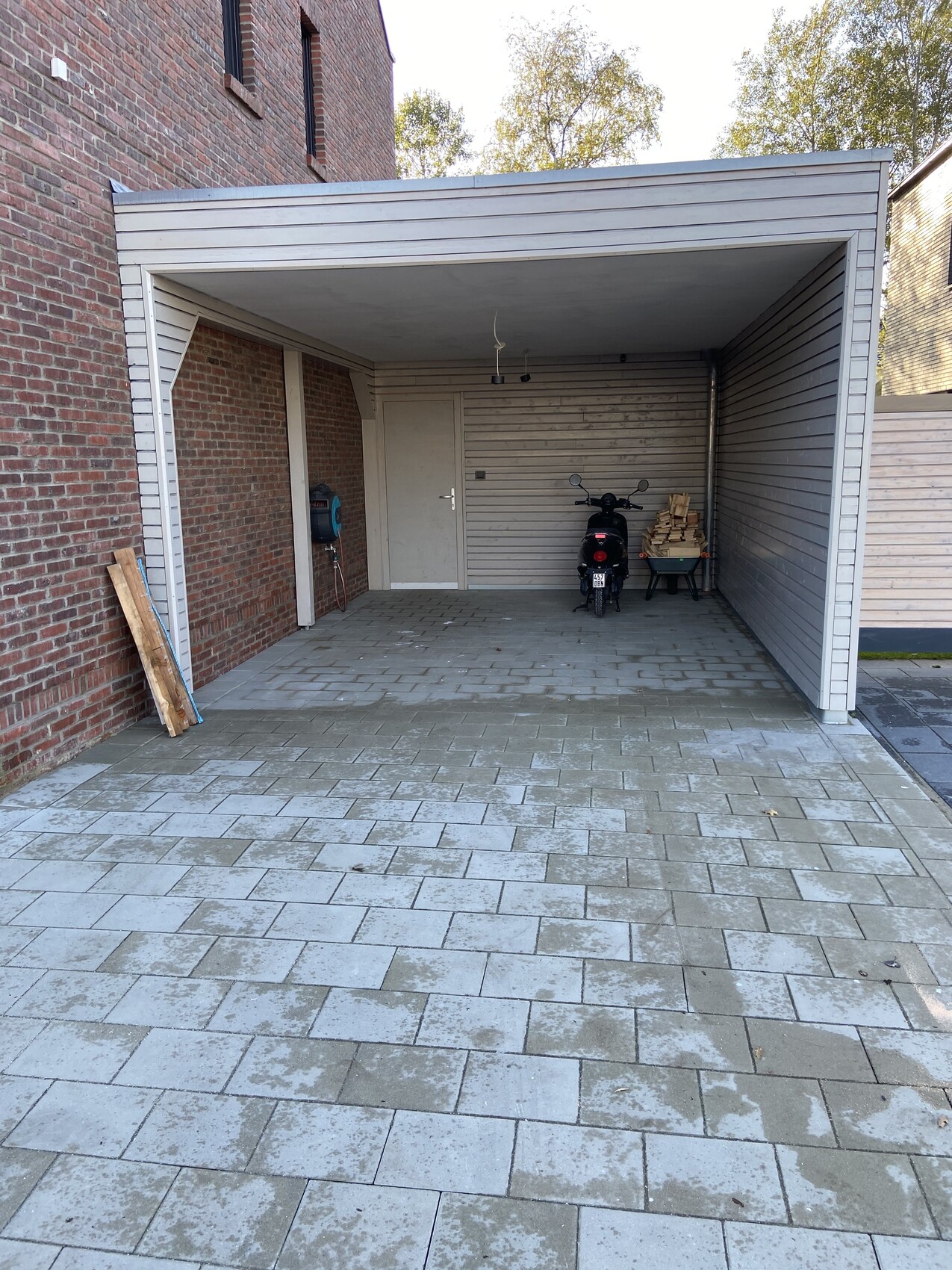 Offener Carport mit Motorrad vor Backstein- und Holzwand, Brennholzstapel rechts, gepflasterter Boden.