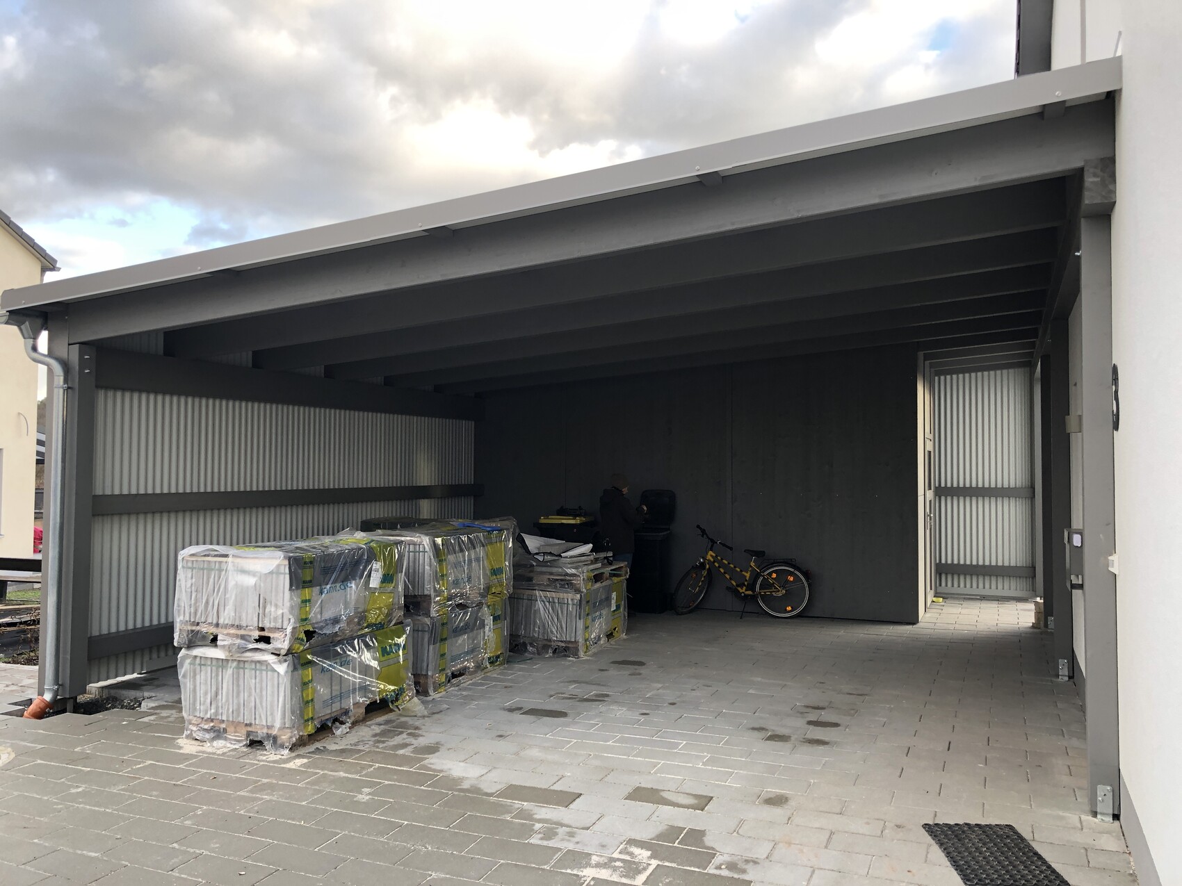 Offener Carport mit gestapelten Paletten links, Fahrrad rechts und Person daneben.