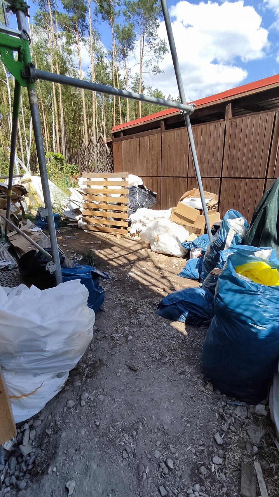 Außengelände mit Müllsäcken, Paletten, Karton und Bauabfällen neben Holzwand; Wald im Hintergrund.